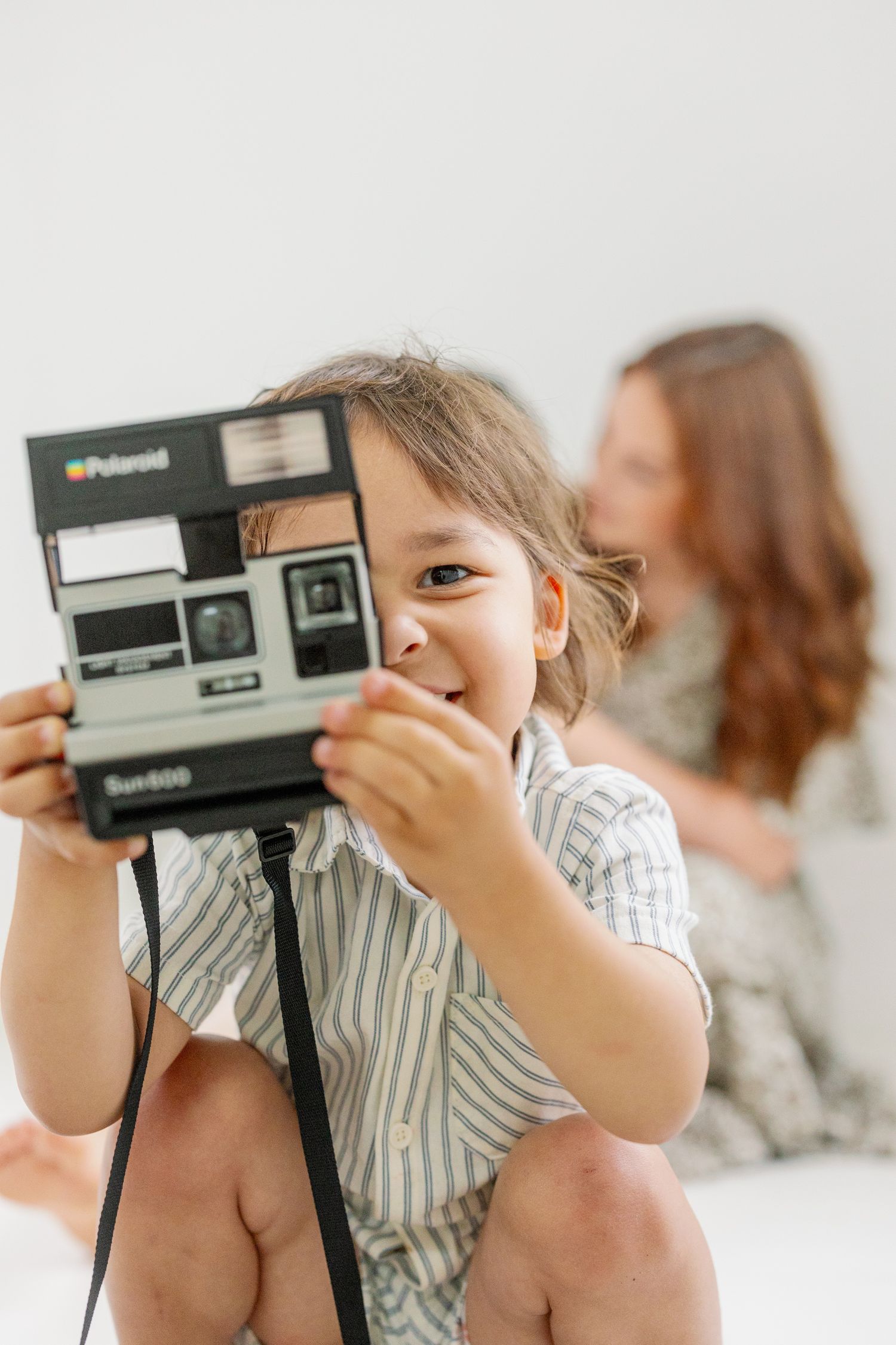 Young child in striped shirt holds vintage Polaroid camera while sitting indoors in natural light.