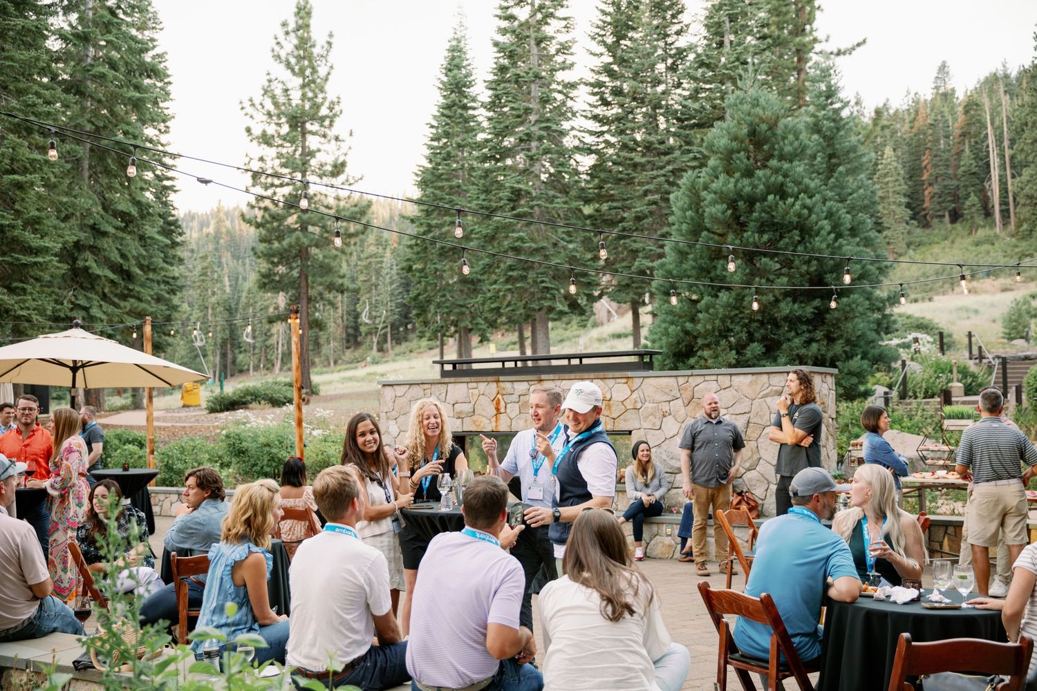 Guests gather at an outdoor mountain venue with towering pine trees during a summer event celebration.