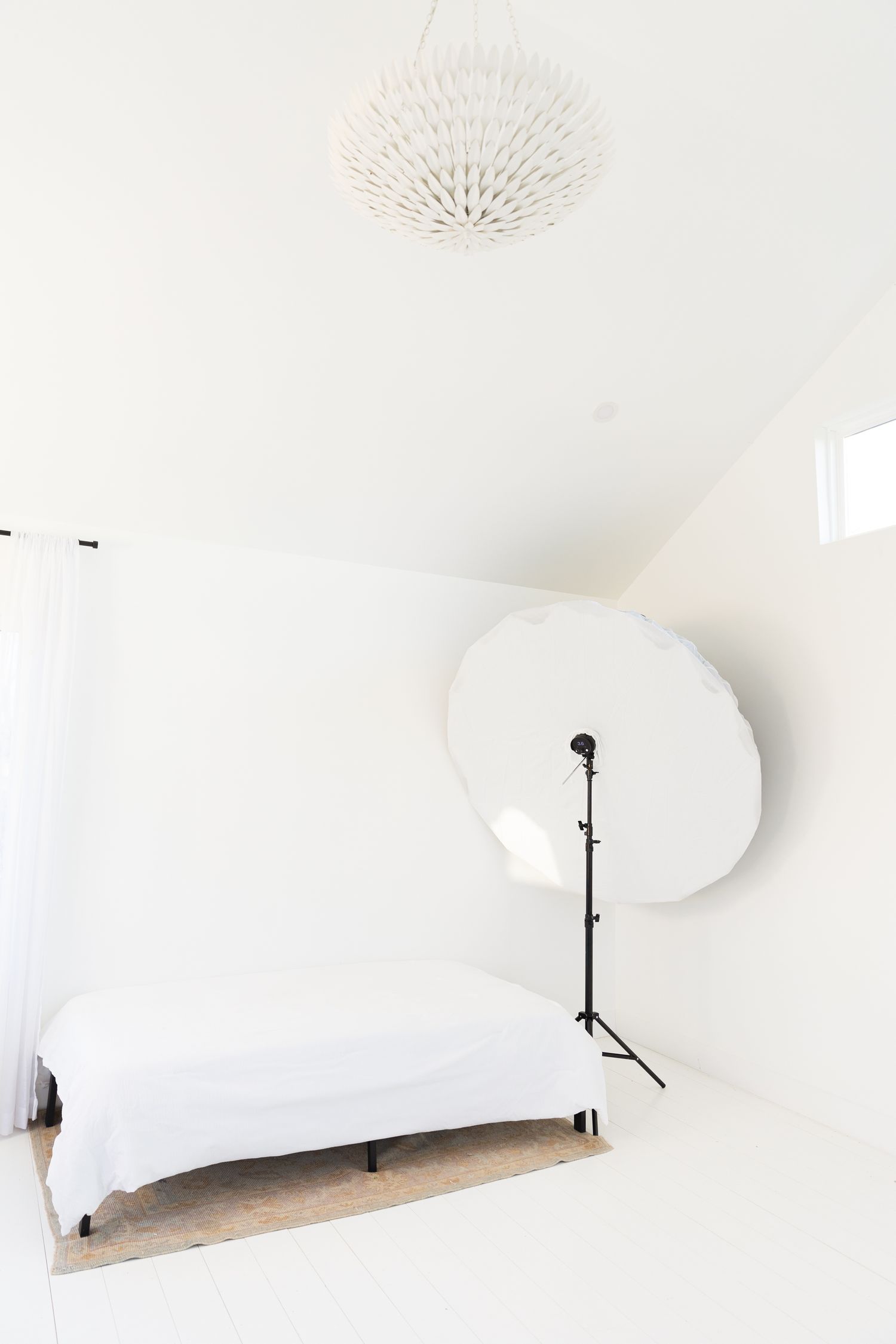 White minimalist photography studio setup with reflector stand and white bedding against clean walls and ceiling.