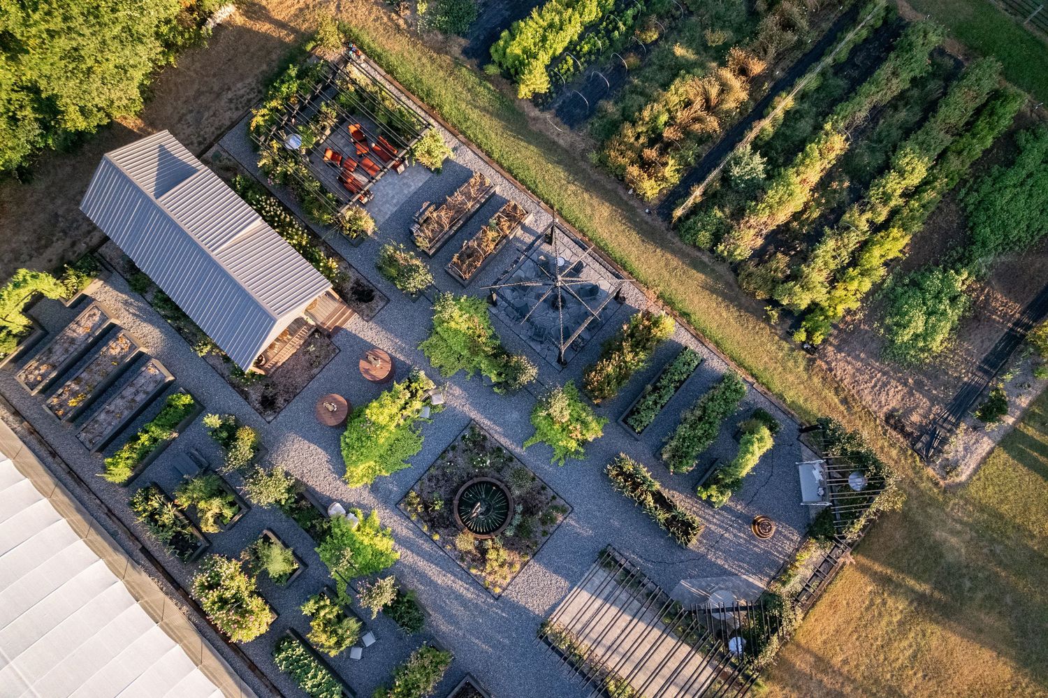 Aerial view of a garden center with greenhouse structures, plant displays, and parking area bathed in warm sunlight.