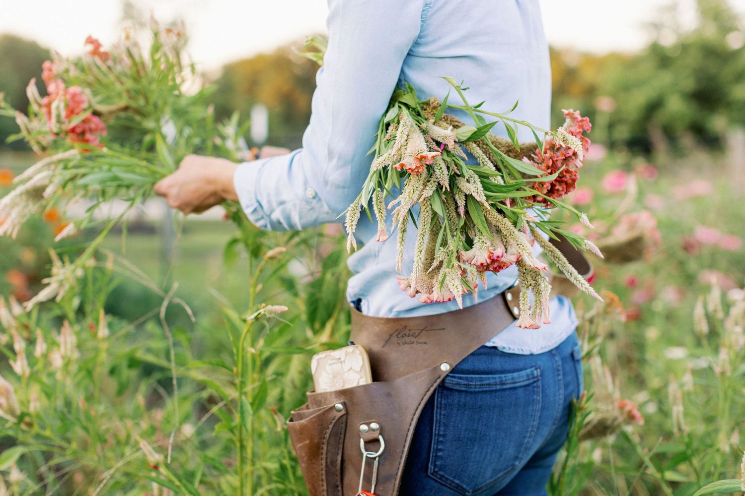Someone holding freshly cut flowers with a brown leather bag at hip level in an outdoor garden setting.