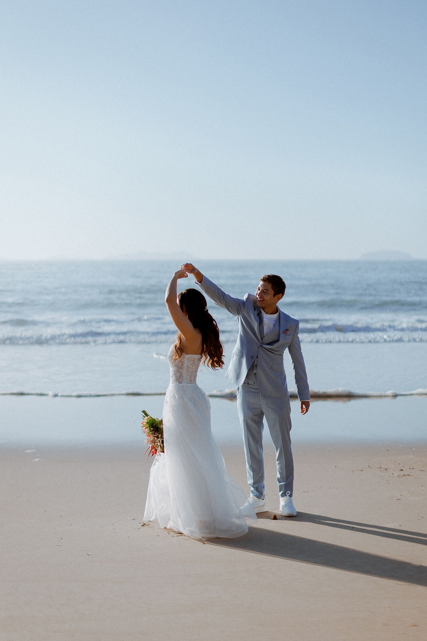 A couple in wedding attire dances together on a sandy beach with ocean waves in the background.