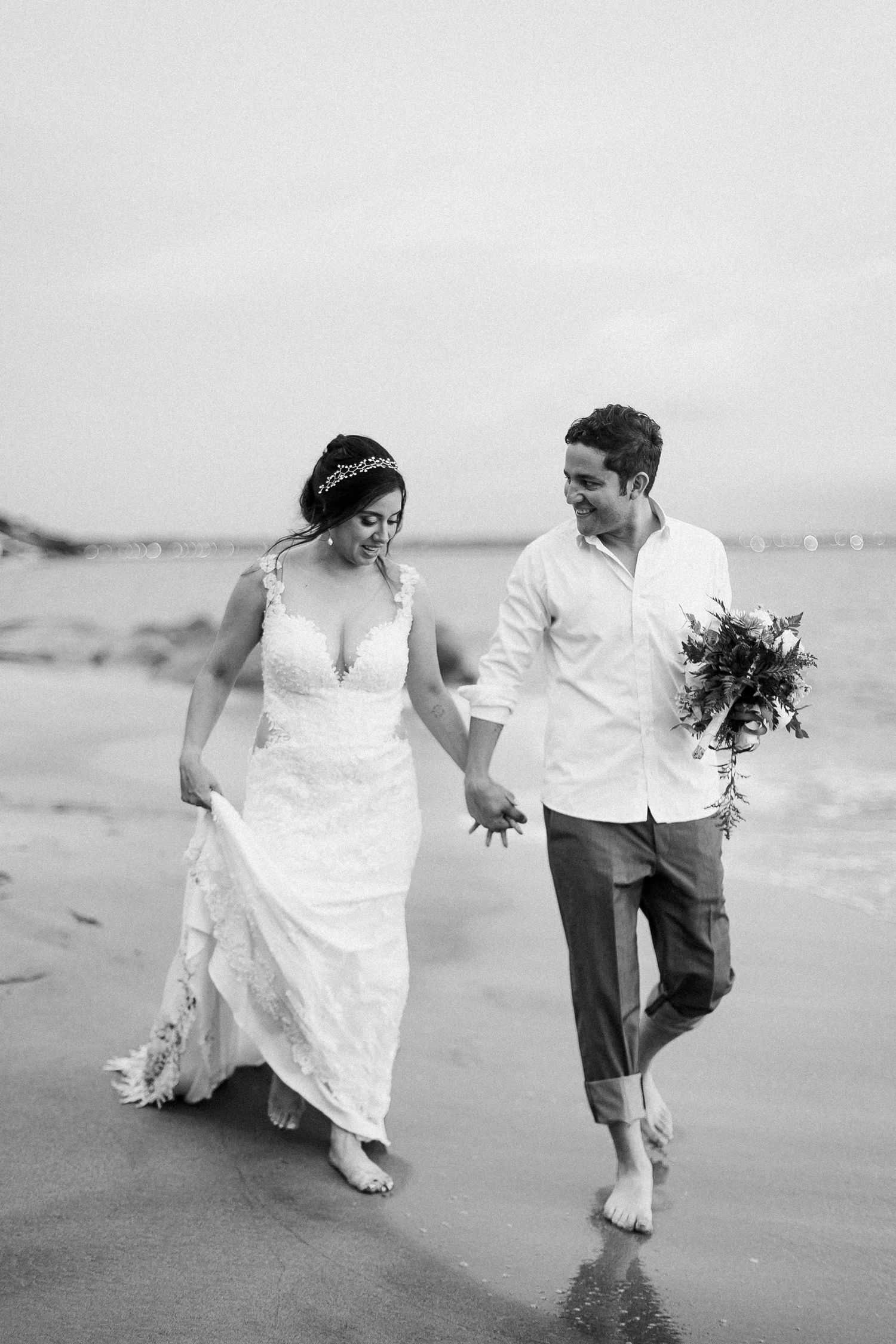 A couple walks hand in hand along a misty beach shoreline in black and white.