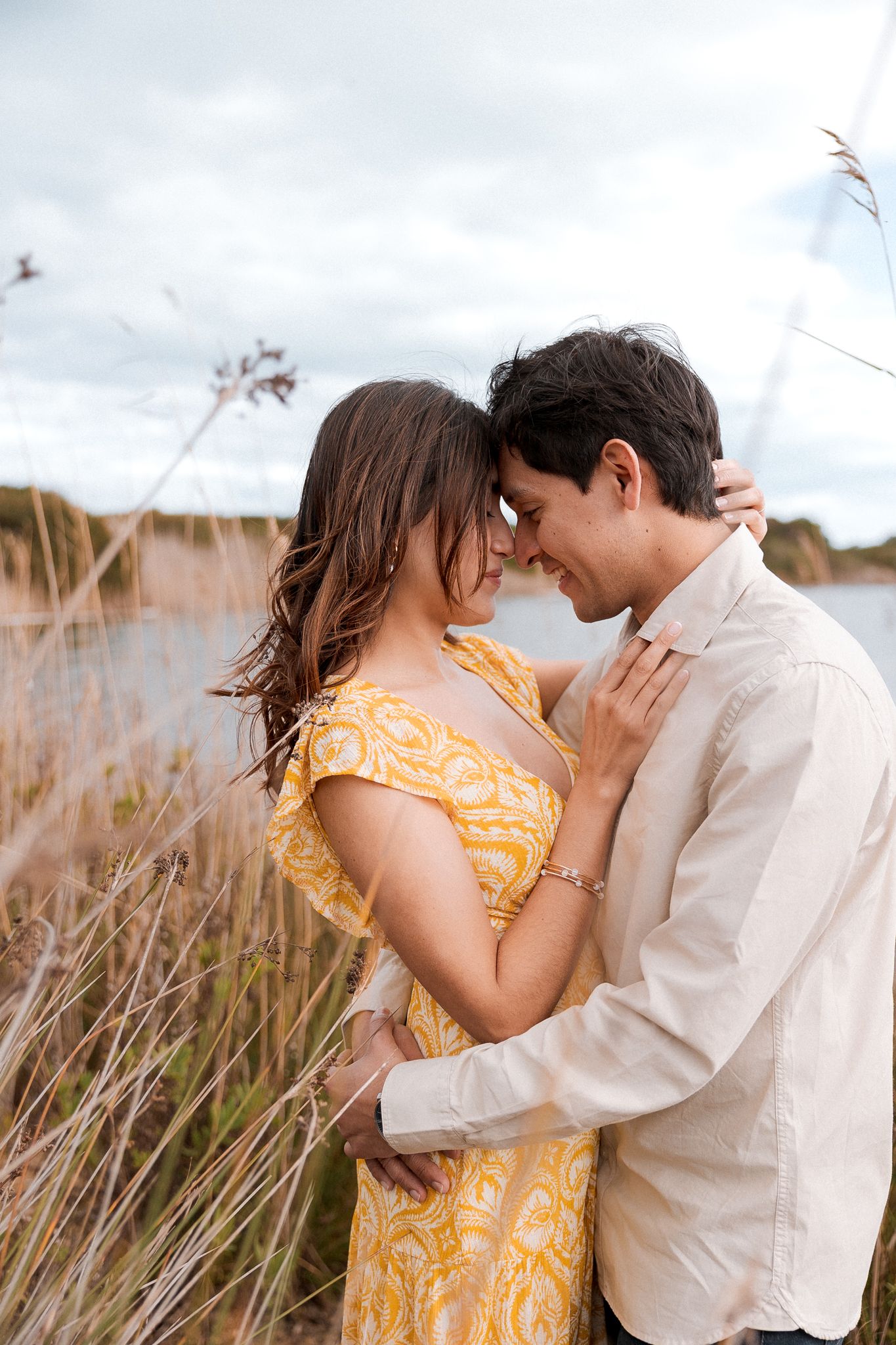 Romantic couple embracing in tall grass with soft lighting and a cloudy sky background.