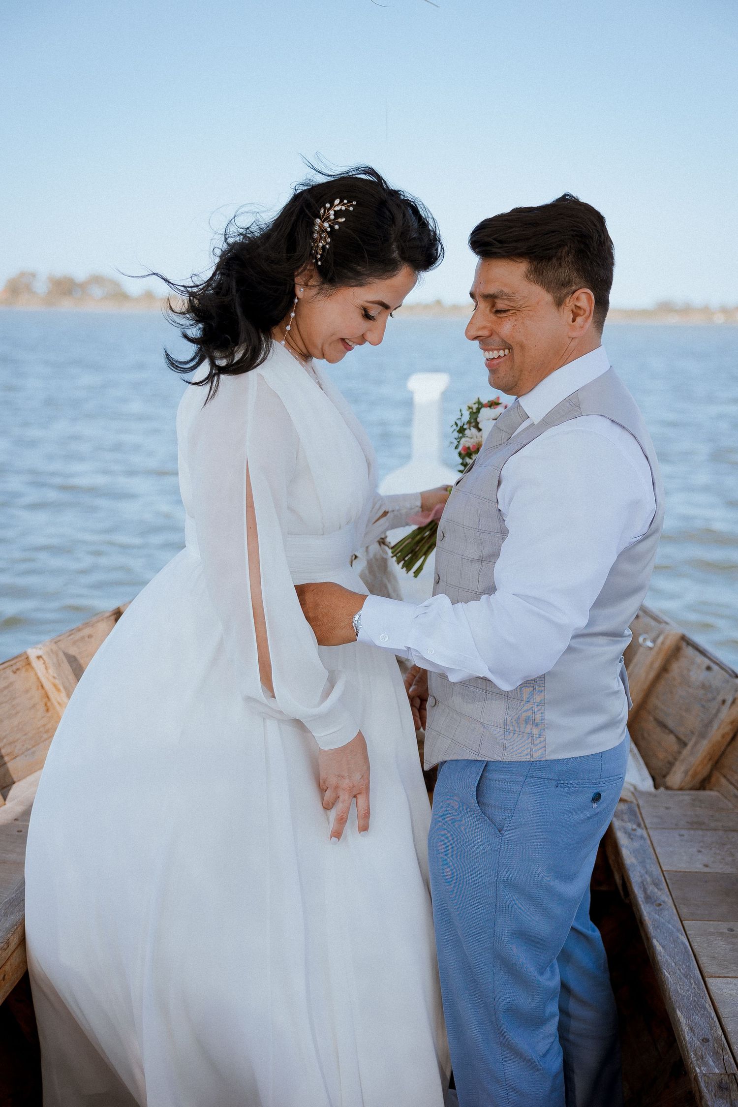 A romantic couple stand together on a wooden boat during their waterfront wedding ceremony.