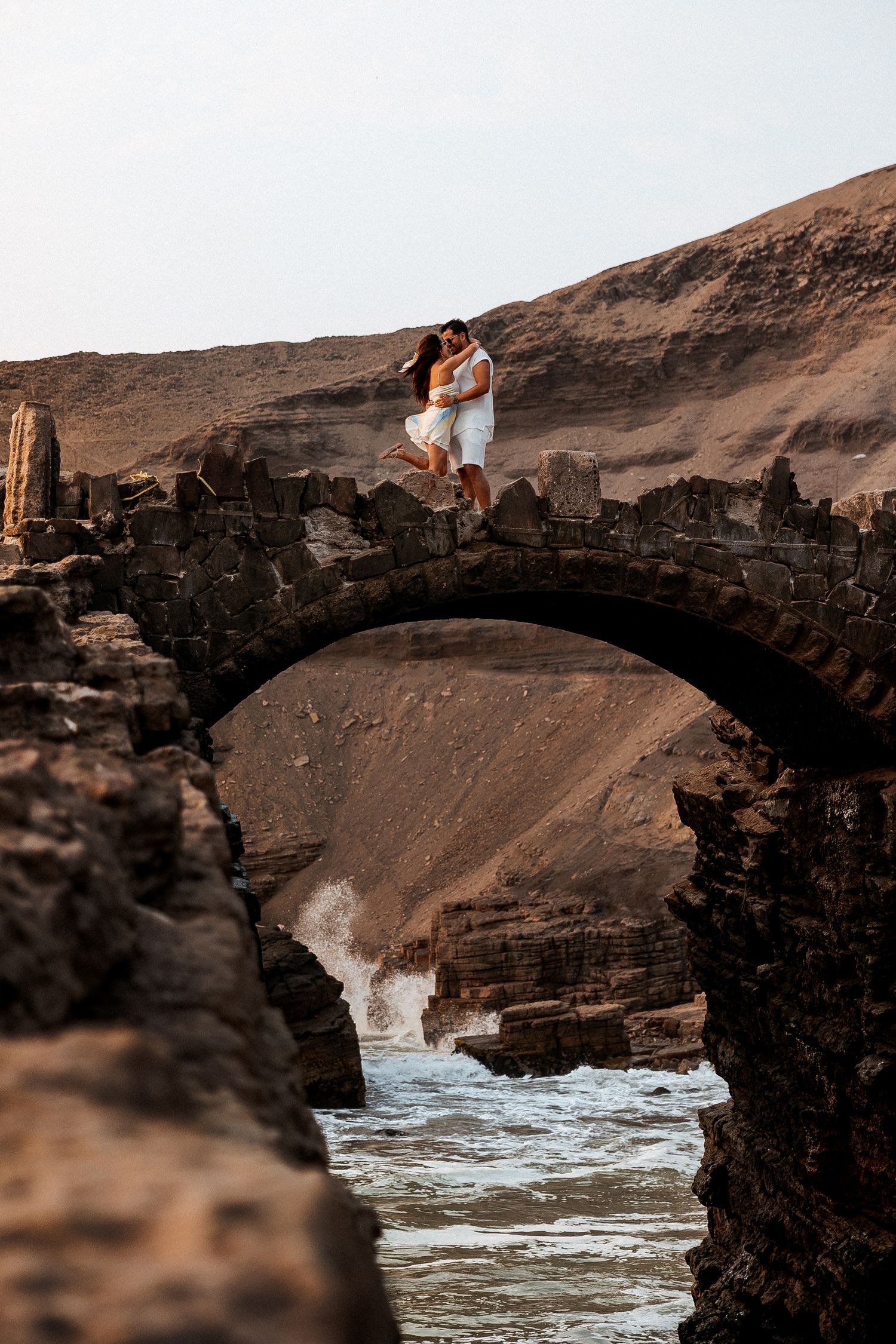 A romantic couple stands on a dramatic stone arch bridge over crashing waves at sunset.