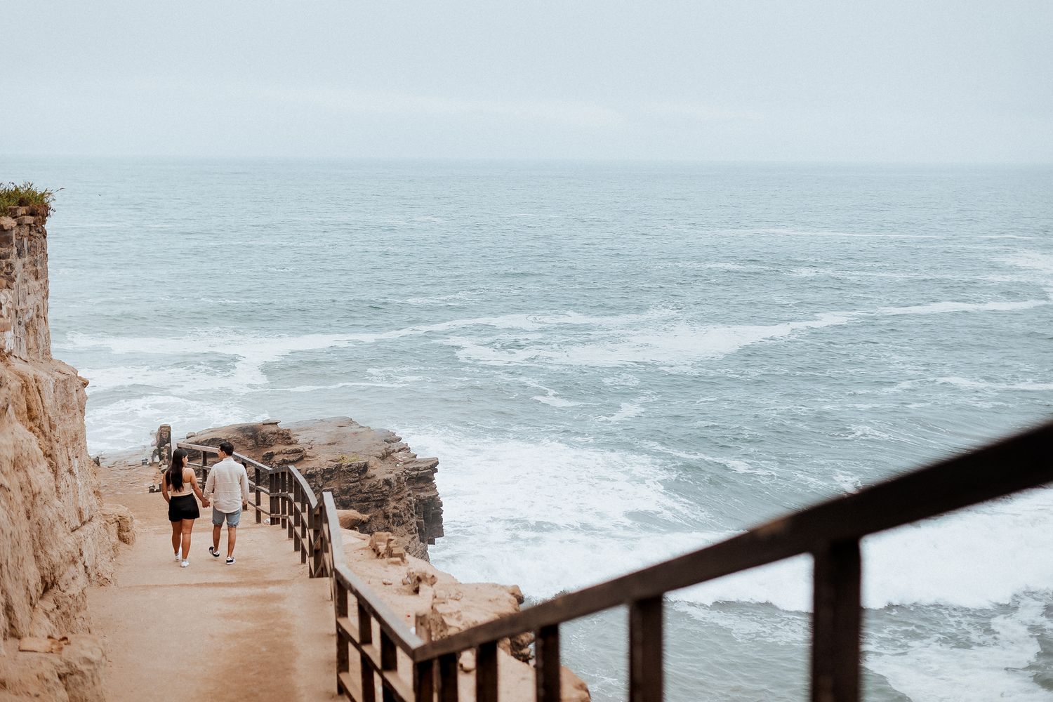Two people walk along a scenic coastal path with railing overlooking rough ocean waves and rocky cliffs on a cloudy day.