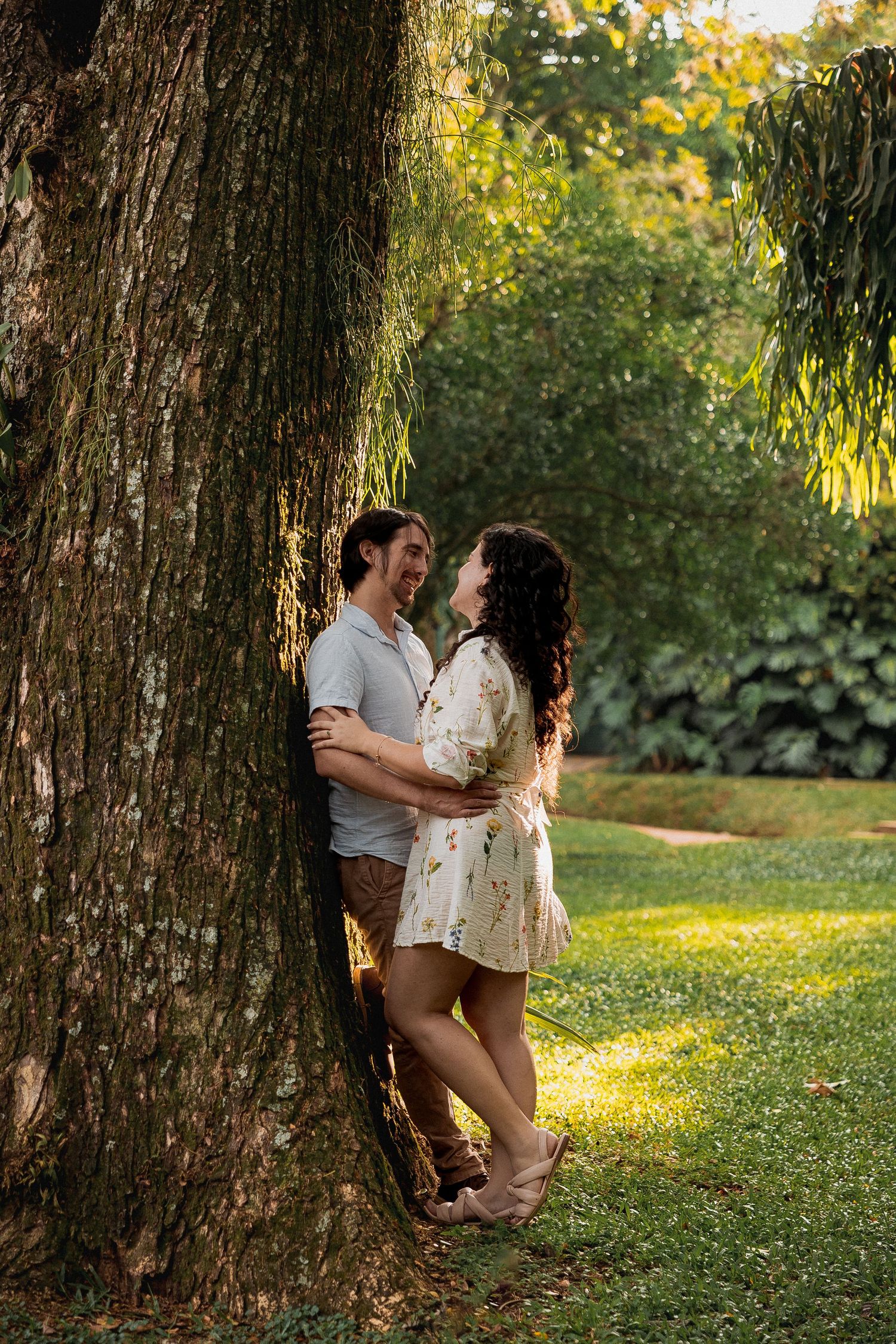 Romantic couple embracing against a large tree trunk in a sunlit garden setting.