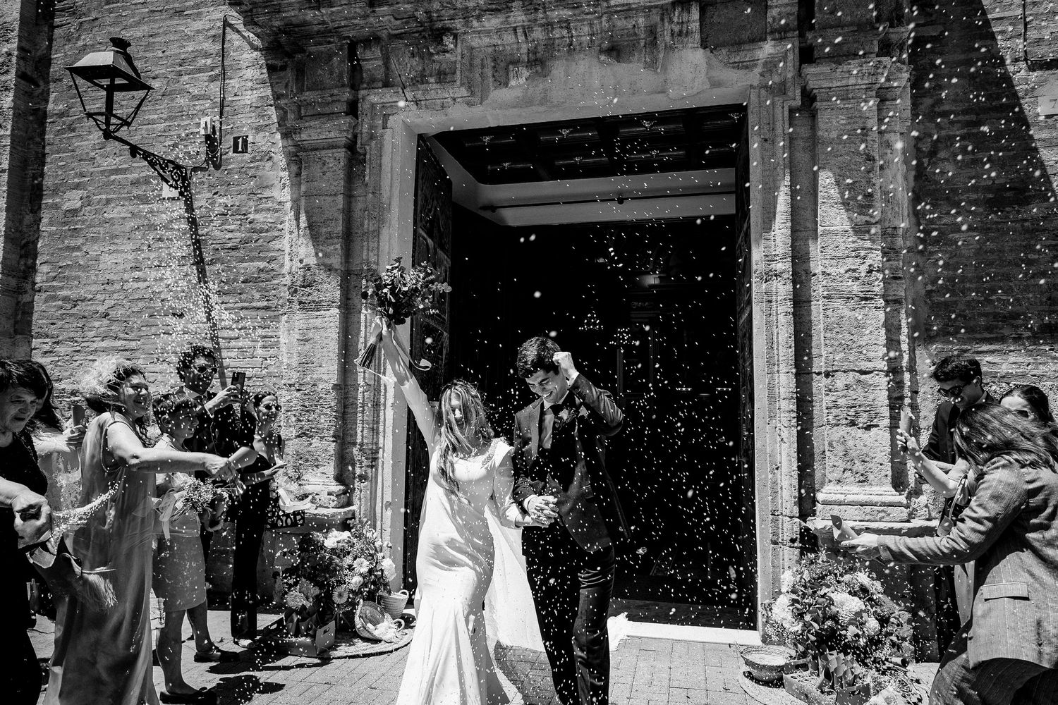 A happy couple exits an old stone church while guests throw confetti during a black and white wedding celebration.