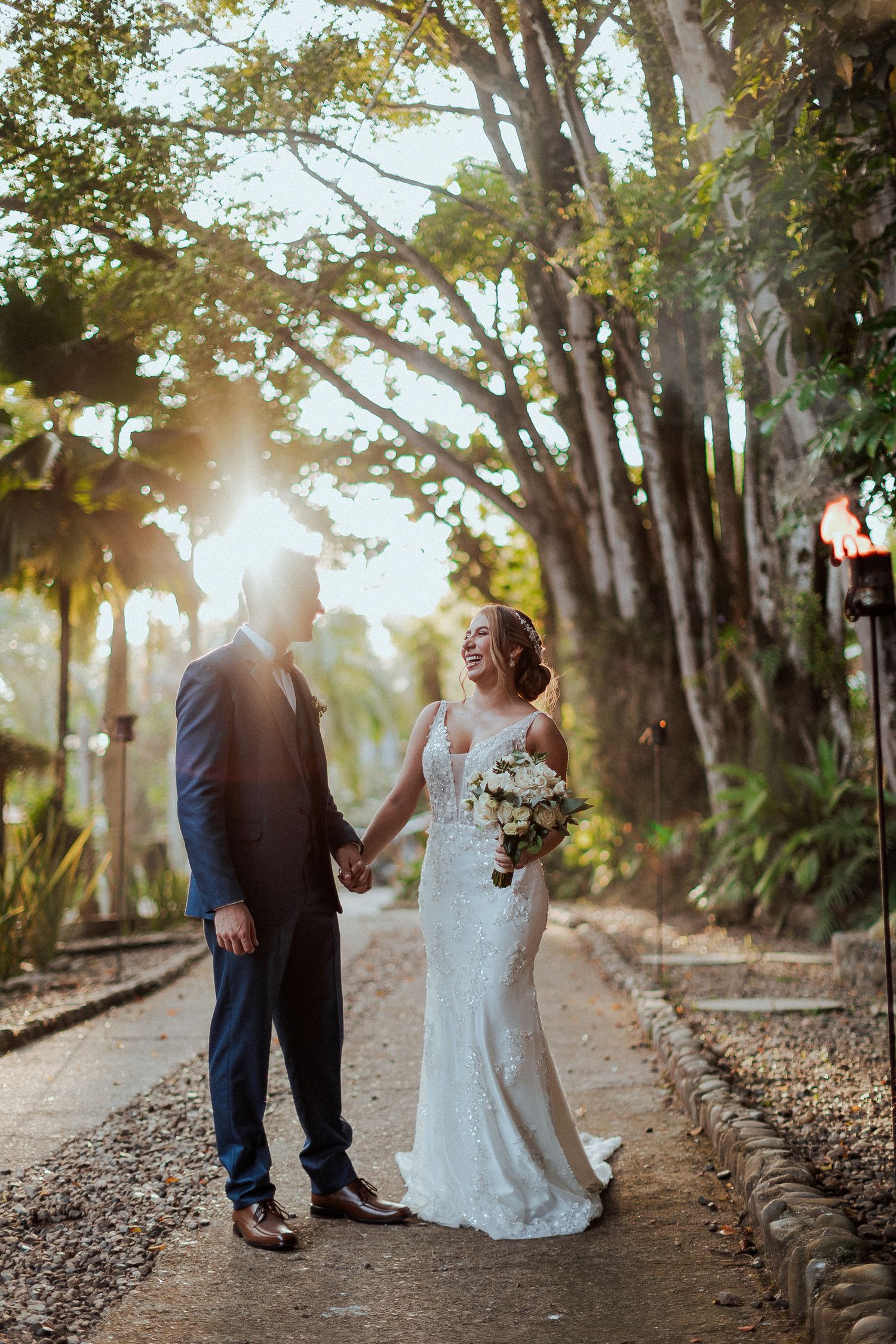 A romantic wedding couple shares a moment on a garden path as sunlight streams through tall trees in the background.