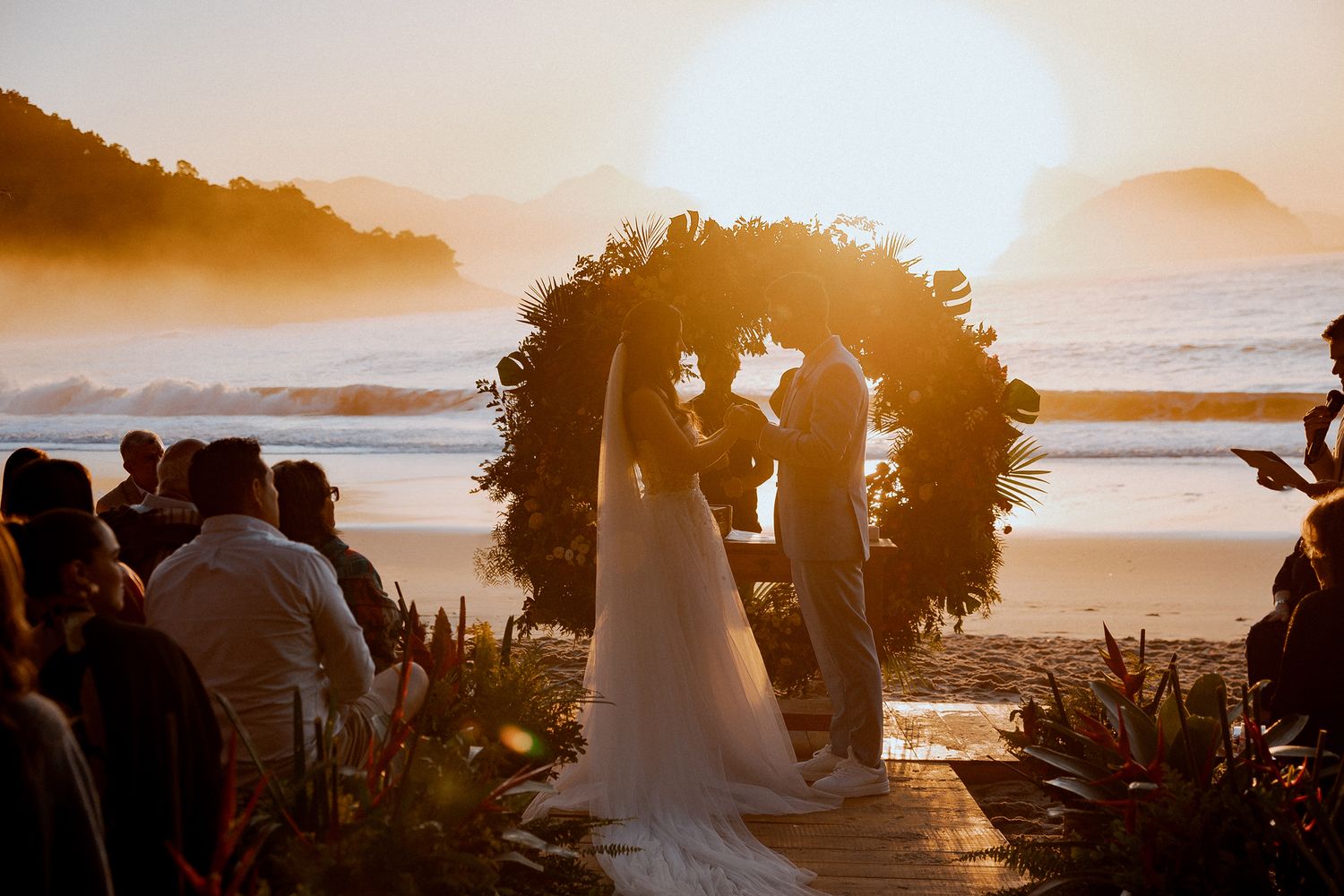 Sunset beach wedding ceremony under floral arch with silhouetted couple and ocean waves in background.