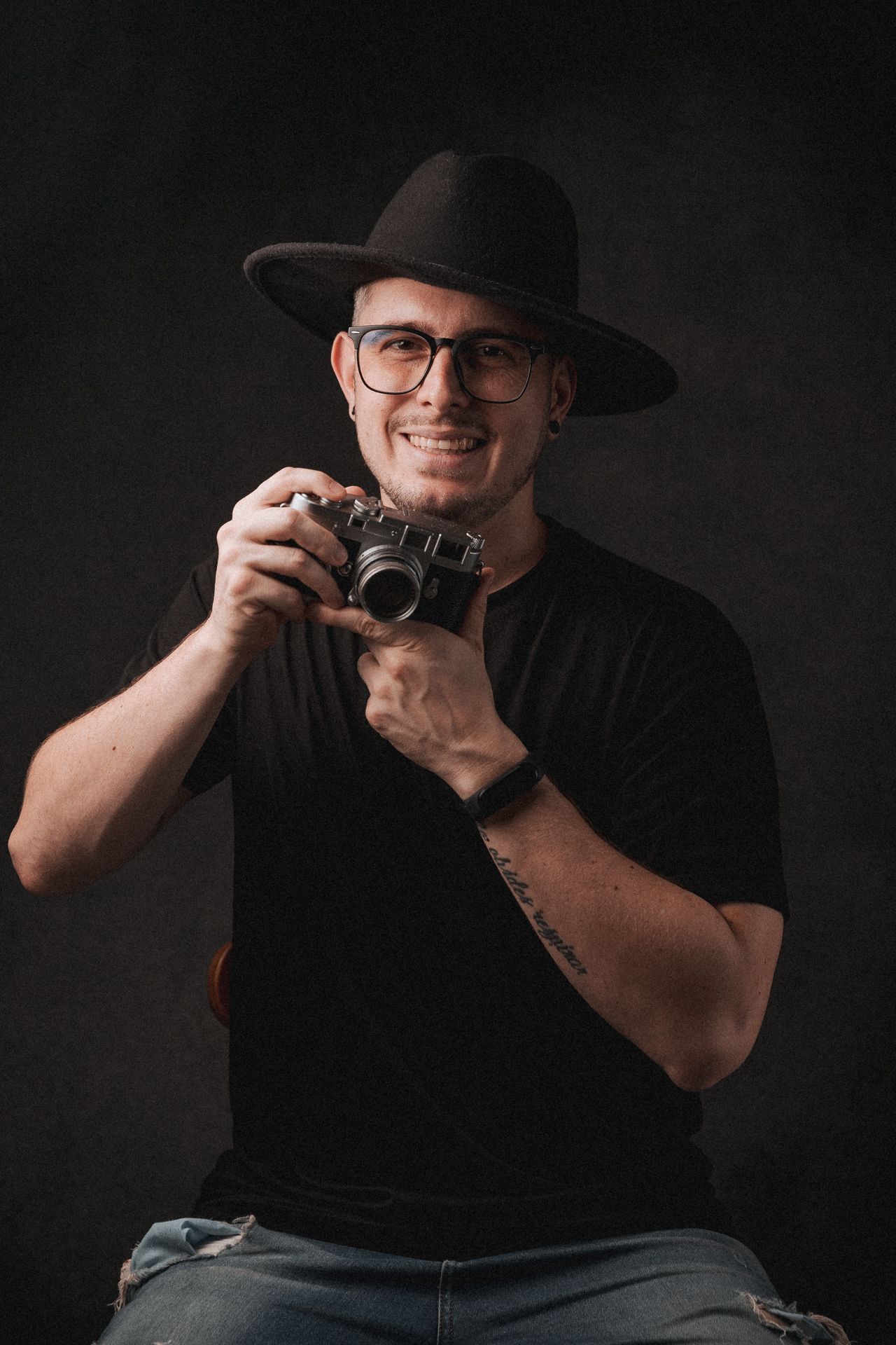 A photographer in black attire and wide-brimmed hat holds a vintage film camera against a dark background.