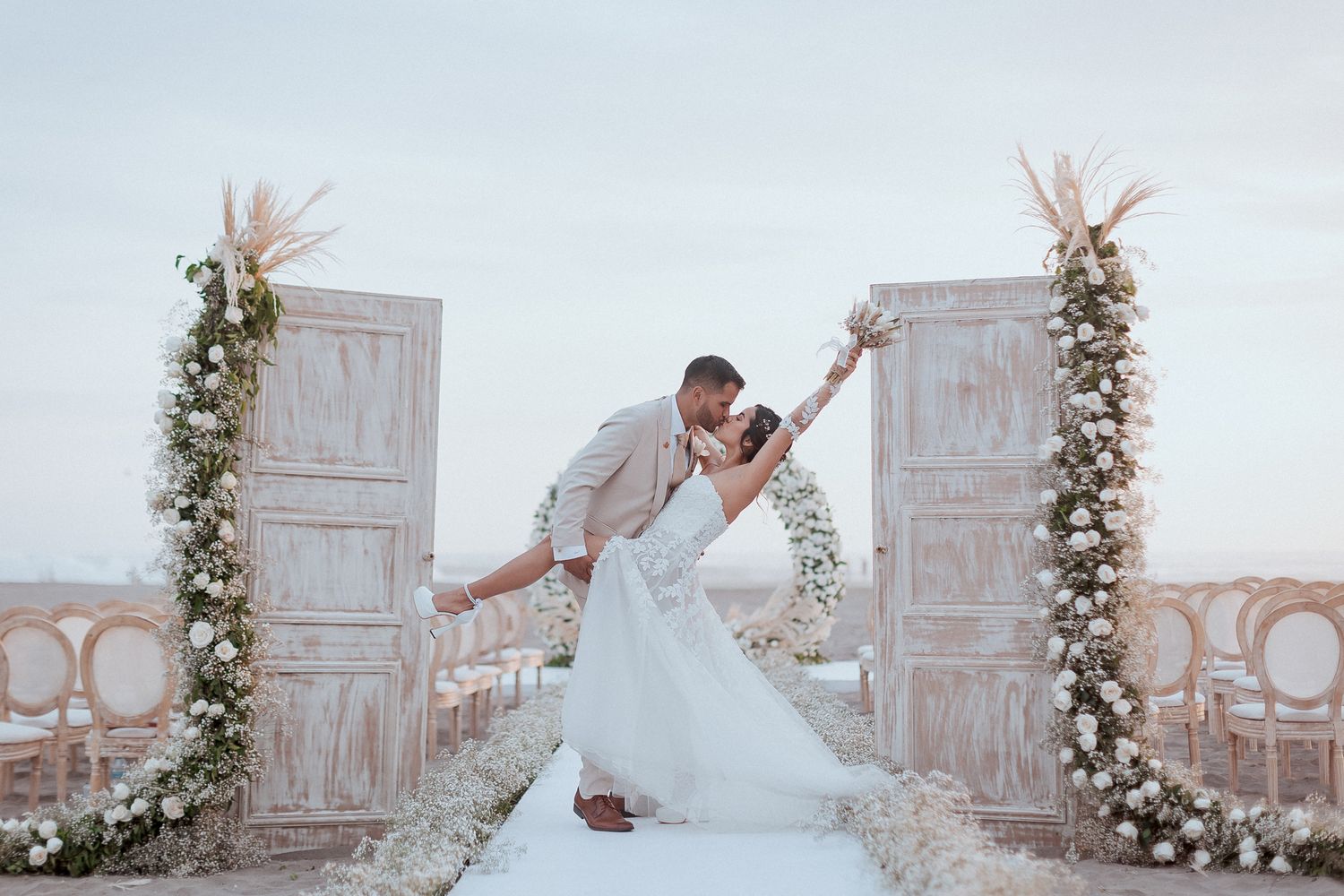 A romantic dip pose between elegant white doors decorated with floral arrangements at a hilltop wedding venue.