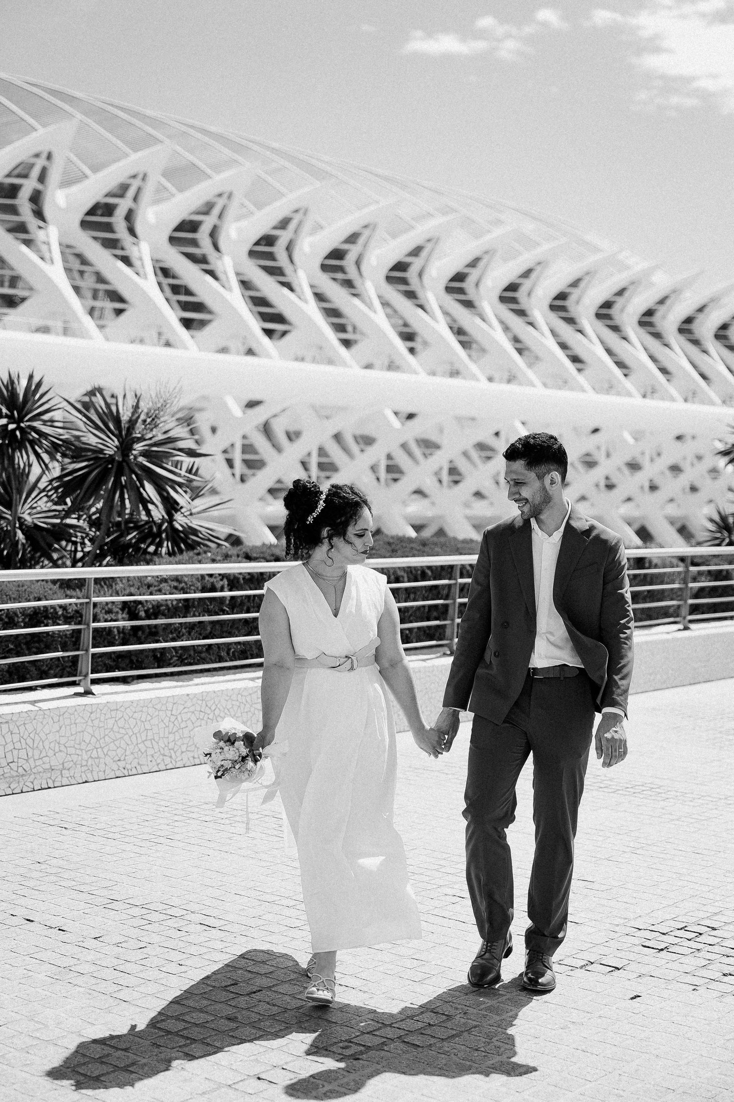 A couple in wedding attire walks hand in hand along a path with the modern architectural structure of Valencia's City of Arts behind them.