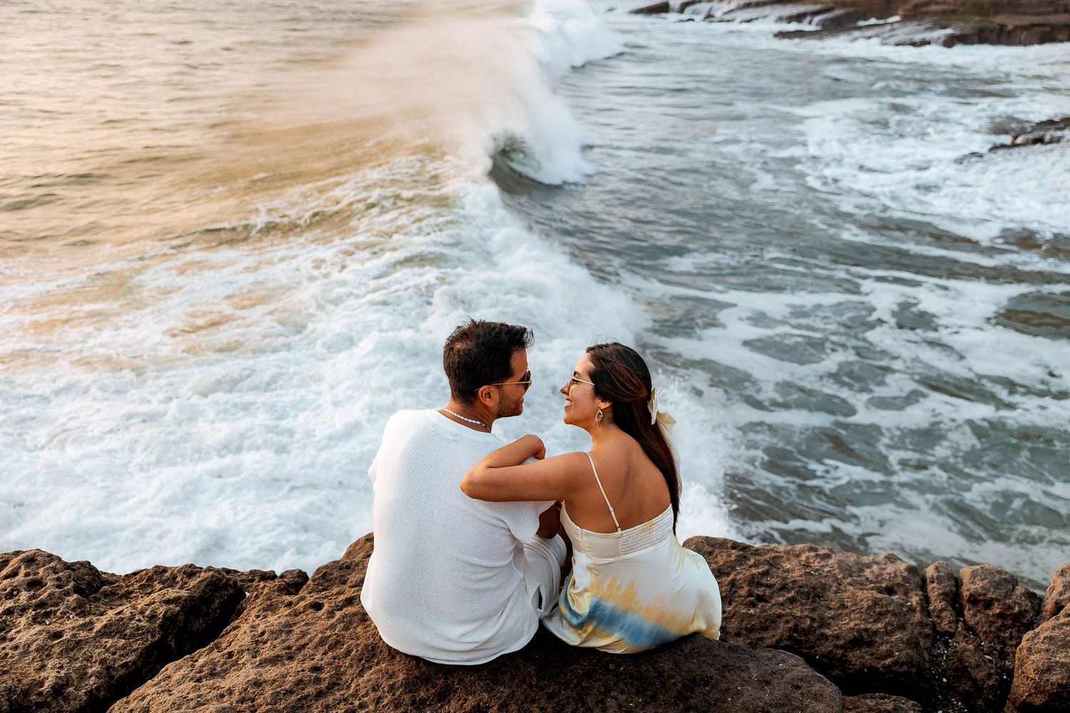 A couple sits on a rocky cliff overlooking dramatic ocean waves crashing against the shore at sunset.