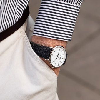 Person wearing a striped sleeve shows their minimalist watch with white face and black leather strap against white clothing.