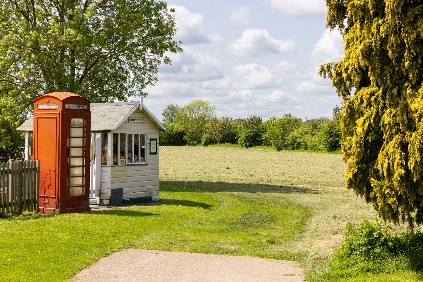 A red British telephone booth stands next to a rustic white building on the edge of a grassy rural field on a sunny day.