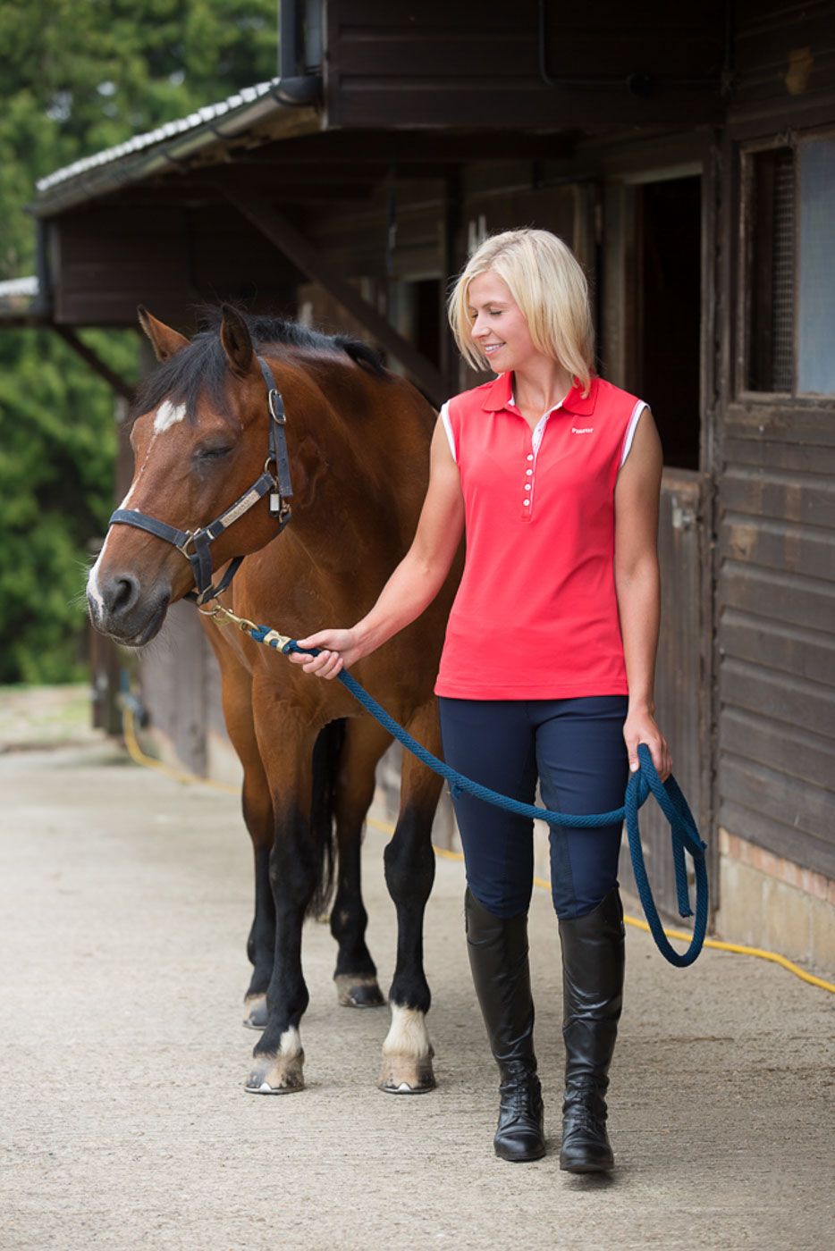 Person in red sleeveless top leads brown horse with black mane outside wooden stable while wearing riding boots.
