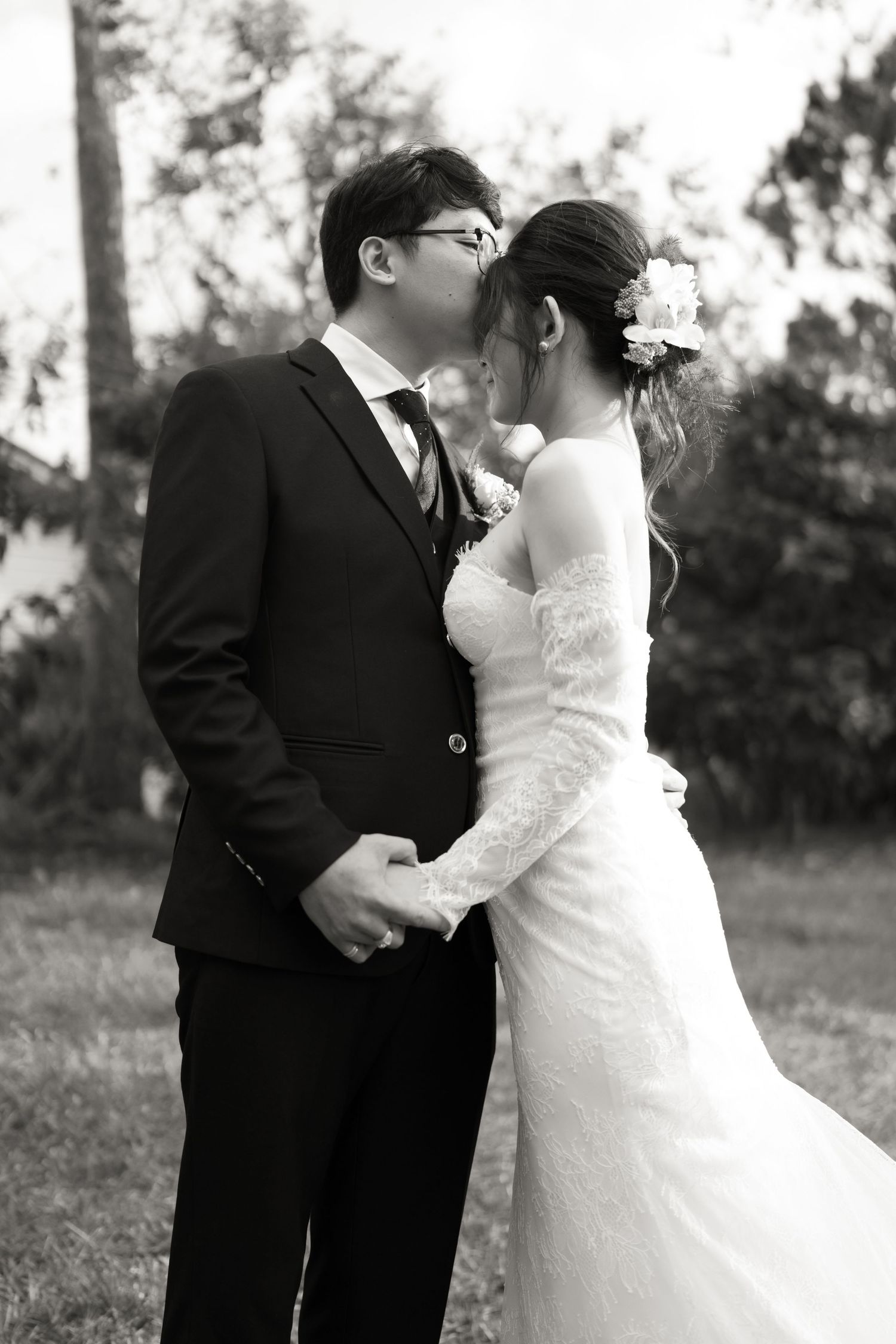 A black and white wedding photo of a couple embracing outdoors, with the bride in a lace dress and groom in dark suit.