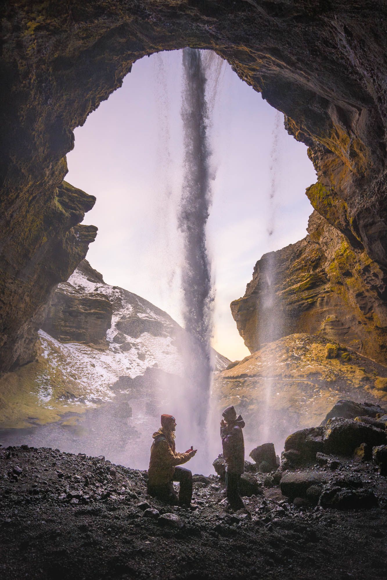 Silhouettes of figures exploring a dramatic cave behind a powerful waterfall with mist and sunlight filtering through.