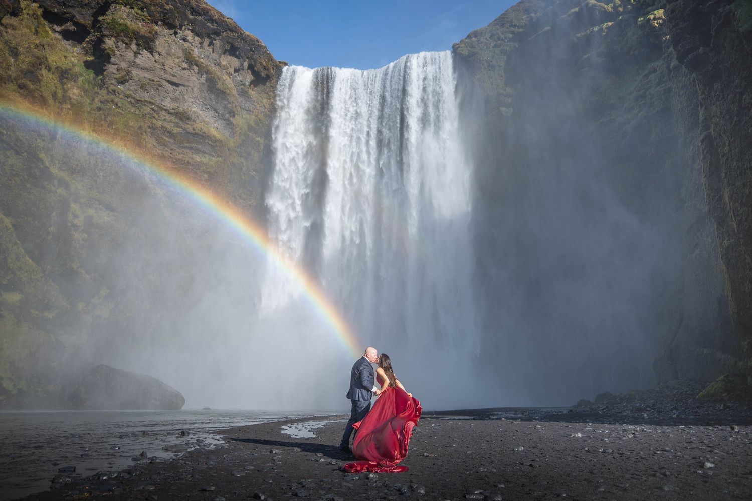 A couple stands before majestic Skogafoss waterfall in Iceland with a vibrant rainbow arcing through the mist.