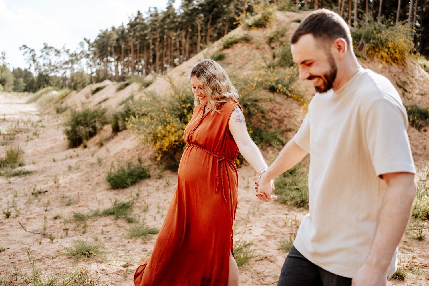 Fröhliches Paar. Werdende Mama in ihrem rostfarbenen Kleid hält lachend die Hand ihres Mannes, während sie barfuß über den Sand gehen.