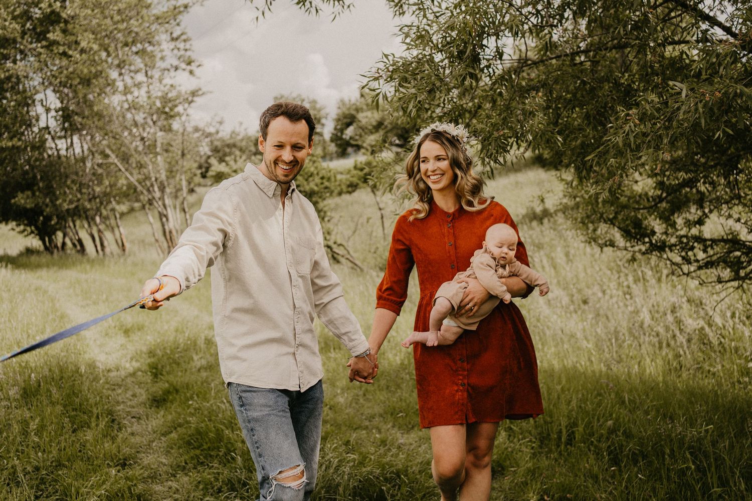 Eltern laufen gemeinsam auf einer Wiese. Mutter hält das Baby im Arm und schaut verliebt zu ihrem Ehemann, beide grinsen glücklich