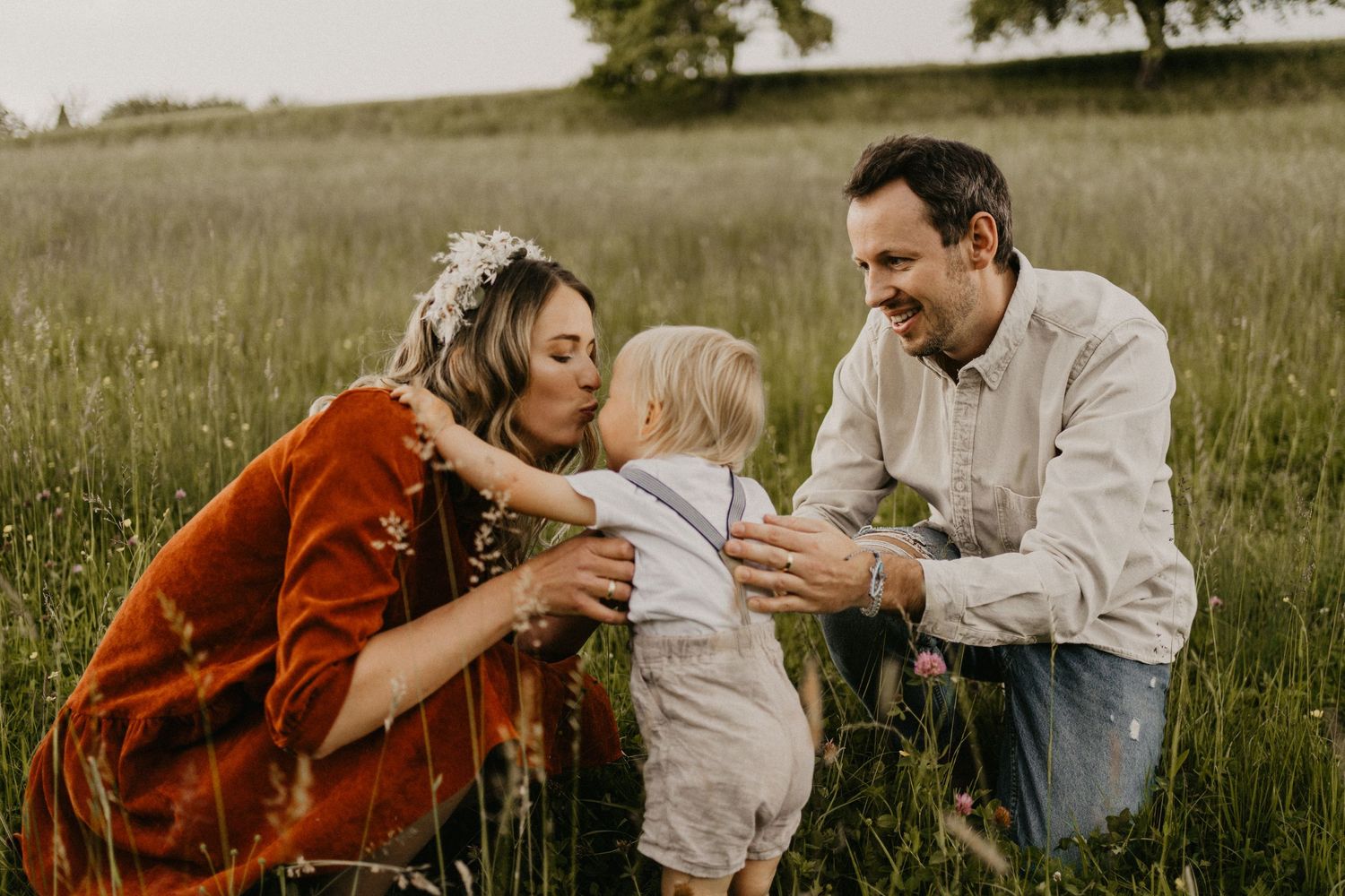 kleiner Junge gibt seiner Mutter einen Kuss in sommerlicher Kulisse, während der Vater liebevoll über den Rücken seines Sohnes streichelt