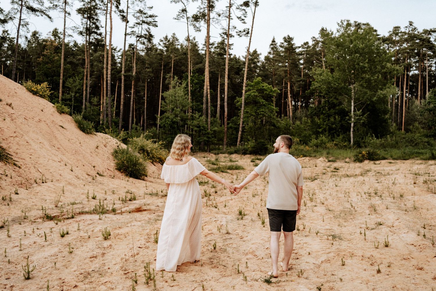 Schwangeres Paar geht barfuß Hand in Hand durch eine natürliche Sandlandschaft bei Roth - Rückenansicht, ruhiger Moment voller Vorfreude.