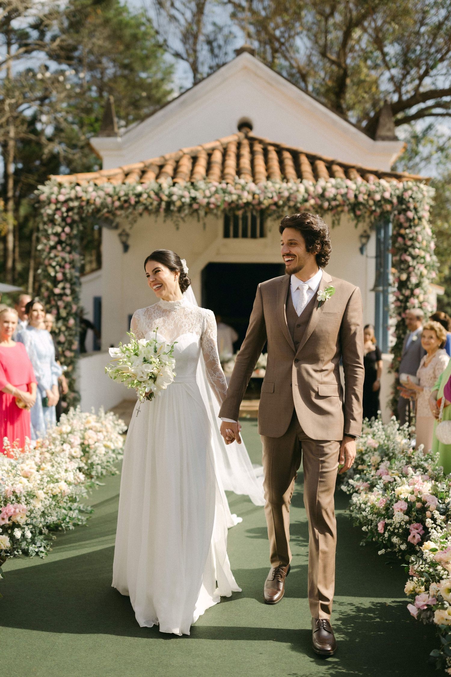A couple walks down a flower-lined aisle under a decorative arch at an outdoor garden wedding ceremony.