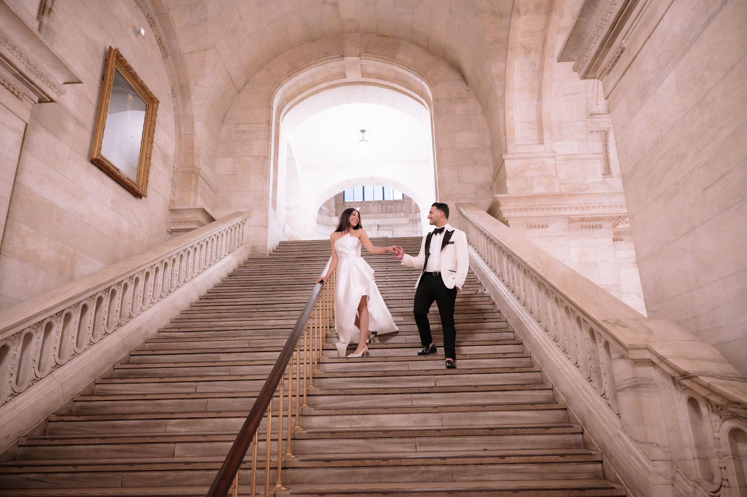A couple in formal attire ascends a grand marble staircase in a historic building.