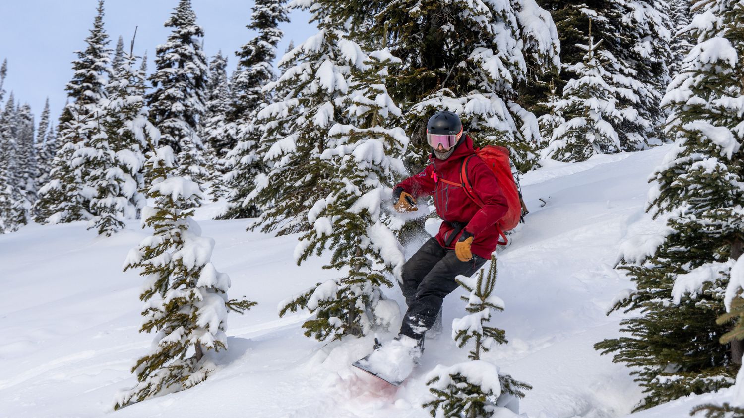 A skier in red outerwear navigates through deep powder snow between snow-laden evergreens.