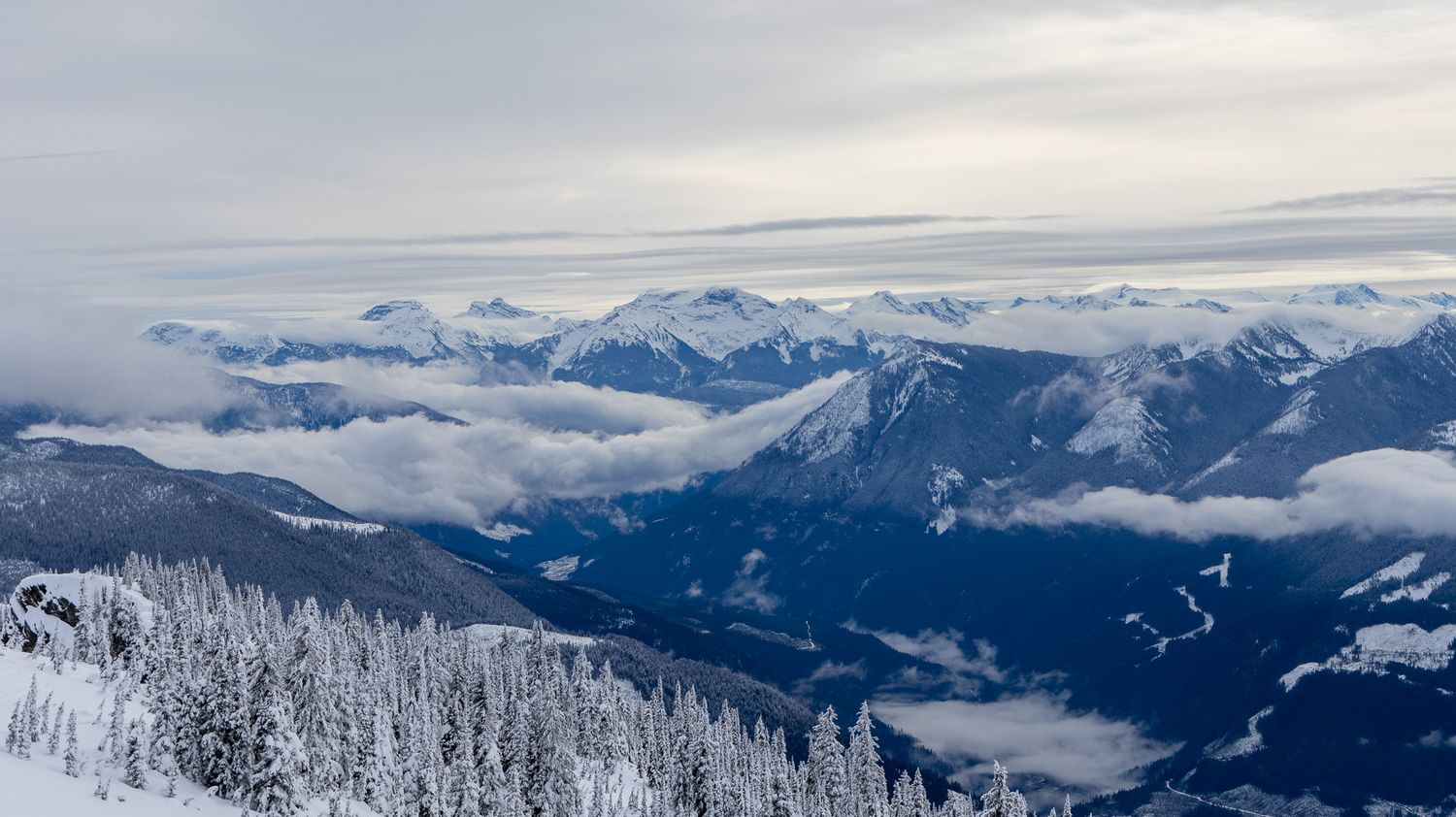 Snow-covered mountain range with low clouds and evergreen forest in winter landscape.