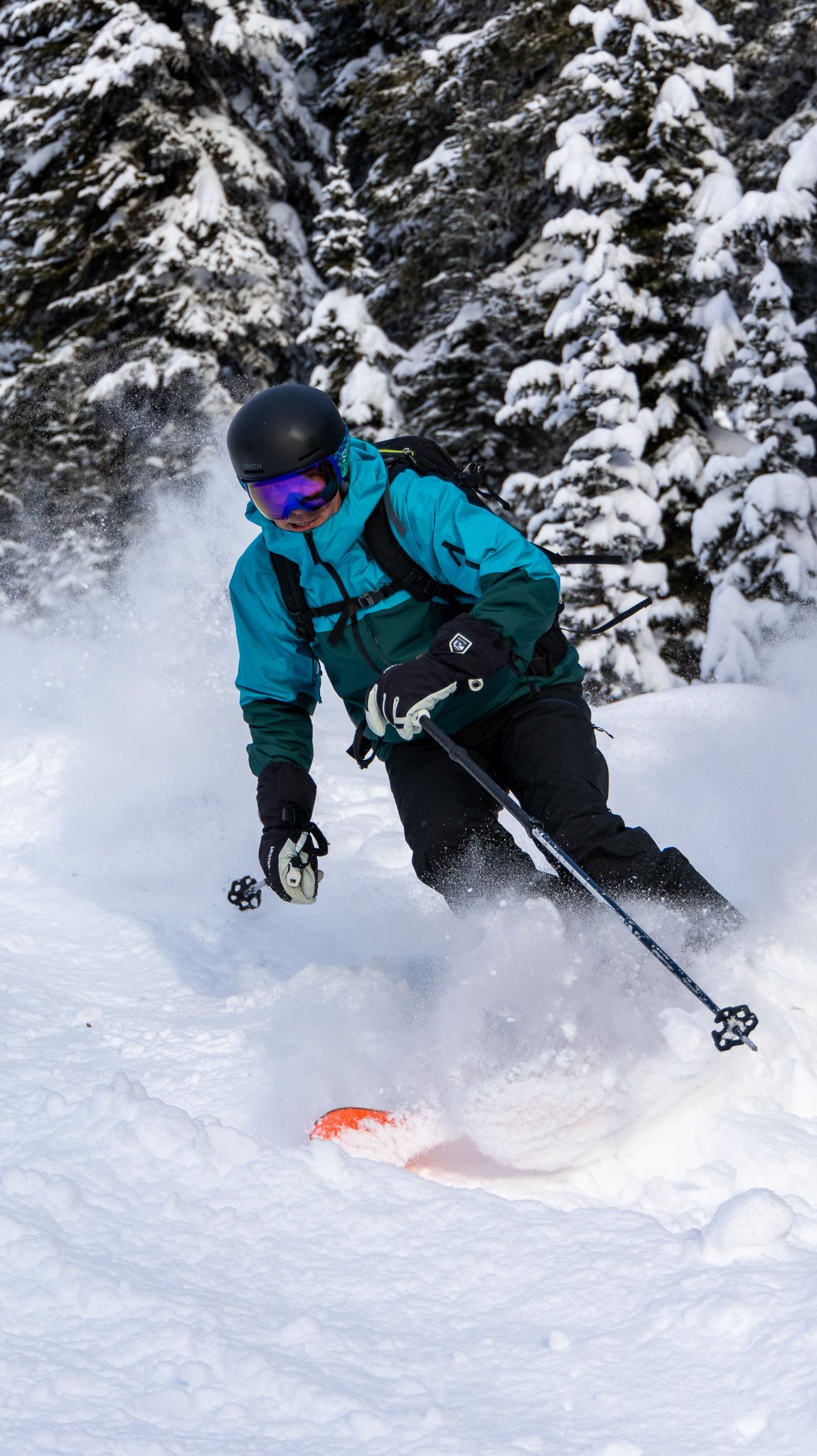 Downhill skier in bright blue jacket kicks up powder snow while descending through snowy forest.