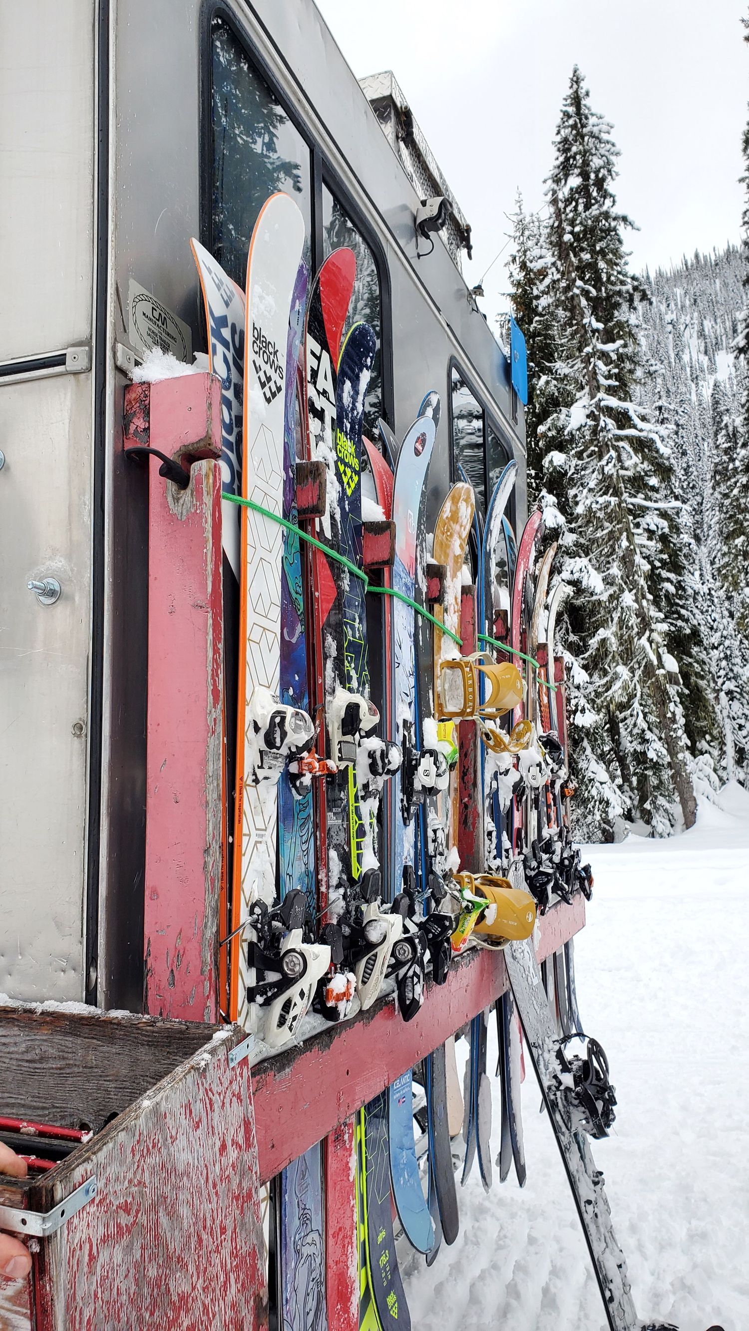 Colorful ski equipment lined up against snowy lodge wall with evergreen trees in background.