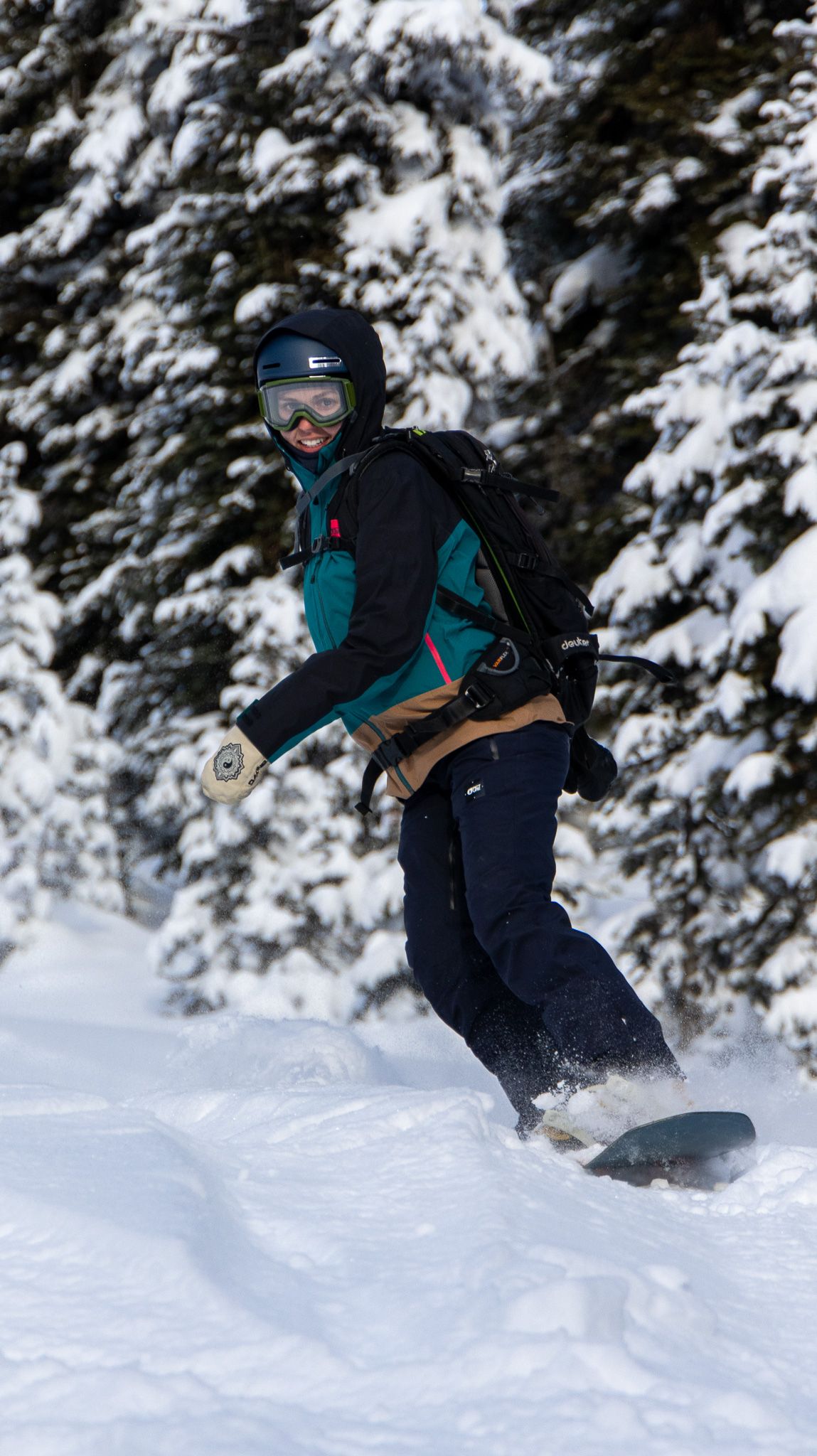 A snowboarder rides through fresh powder snow against a backdrop of snow-covered pine trees.