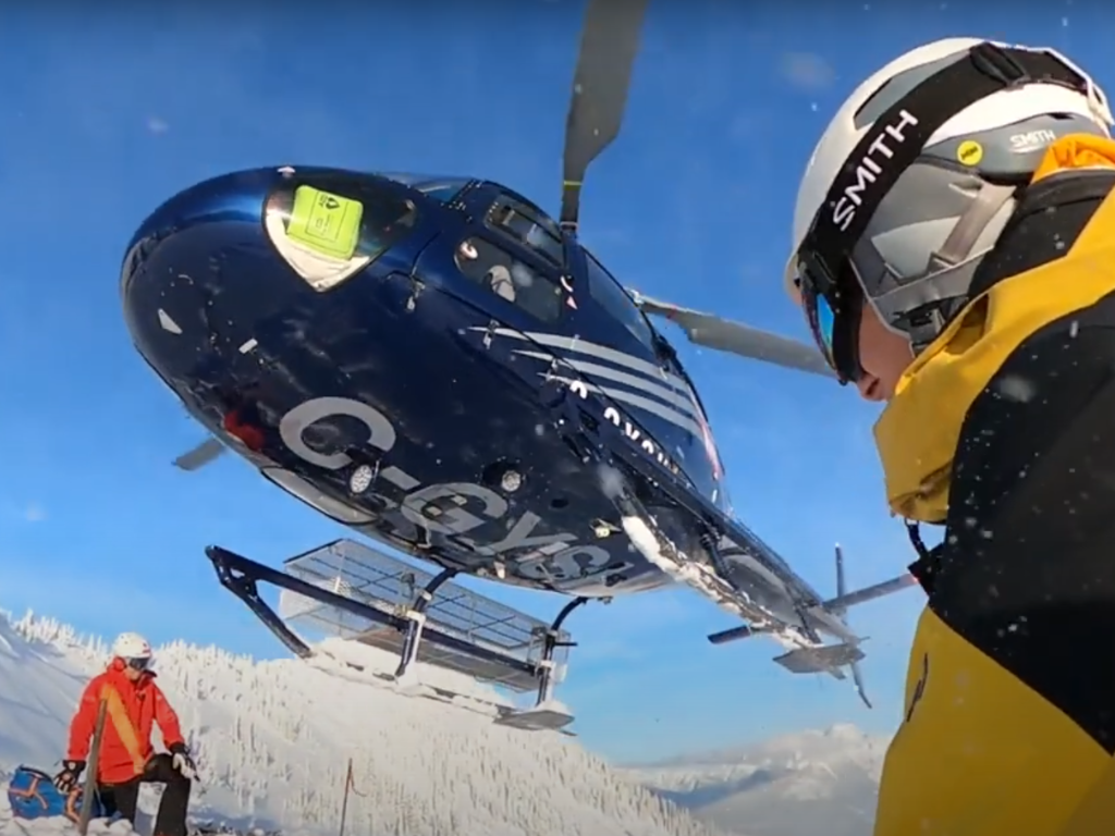 Blue helicopter preparing for takeoff in snowy mountain terrain with skier in foreground.