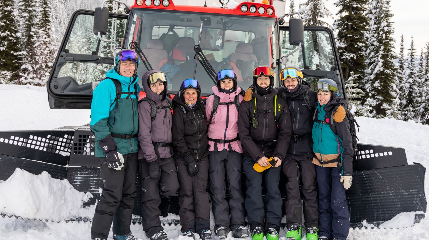 A group poses in winter gear in front of a red snow grooming machine in a snowy mountain setting.