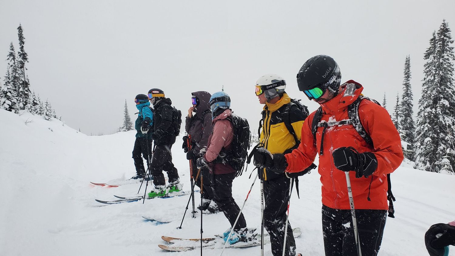 Winter backcountry ski group gathers on snowy slope surrounded by mountain terrain.