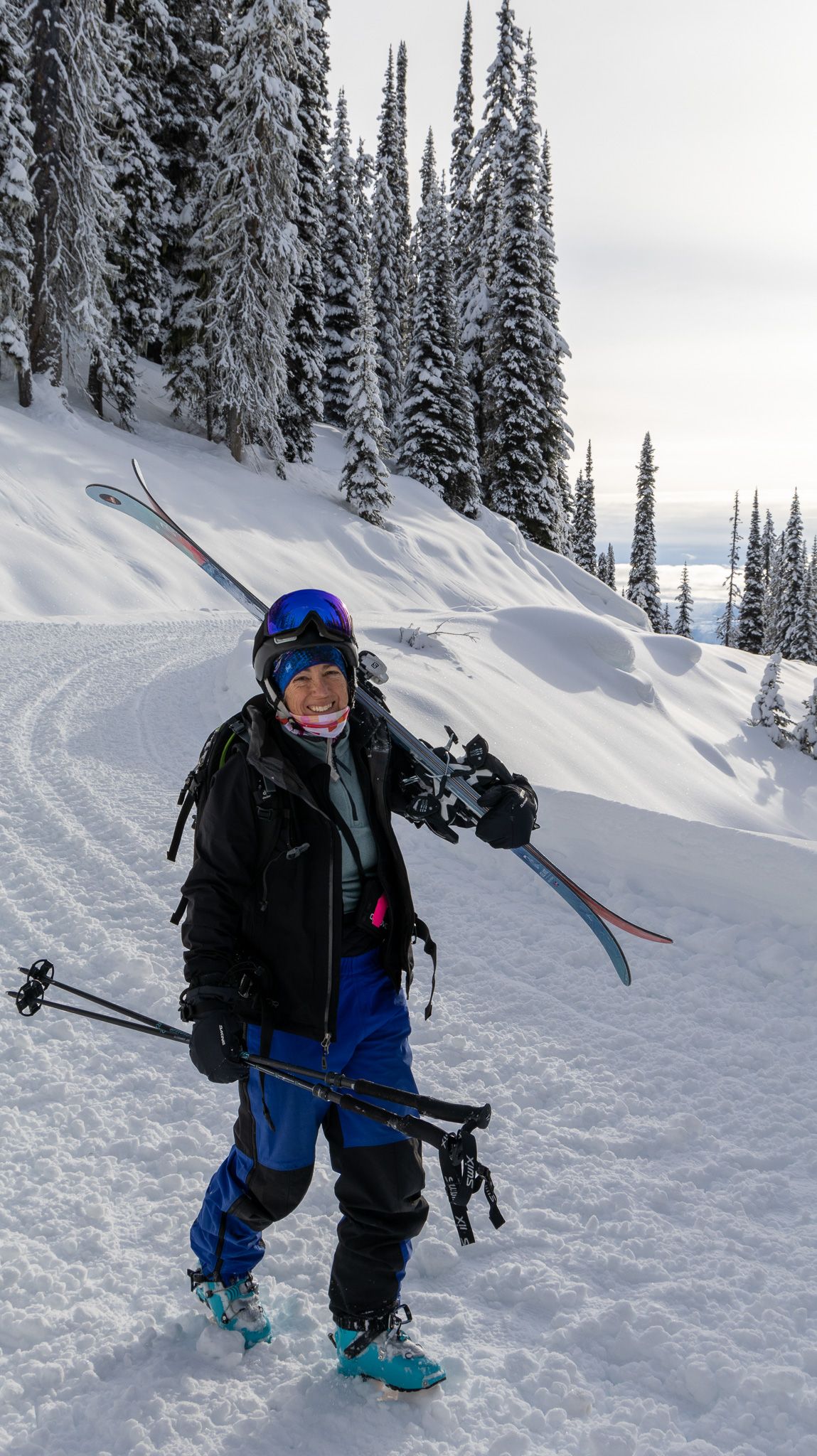 A skier in winter gear stands on a snowy trail with snow-covered evergreens in the background.