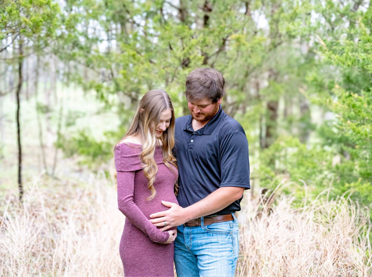 A couple stands close together in a romantic embrace surrounded by tall grass and trees in a natural outdoor setting.
