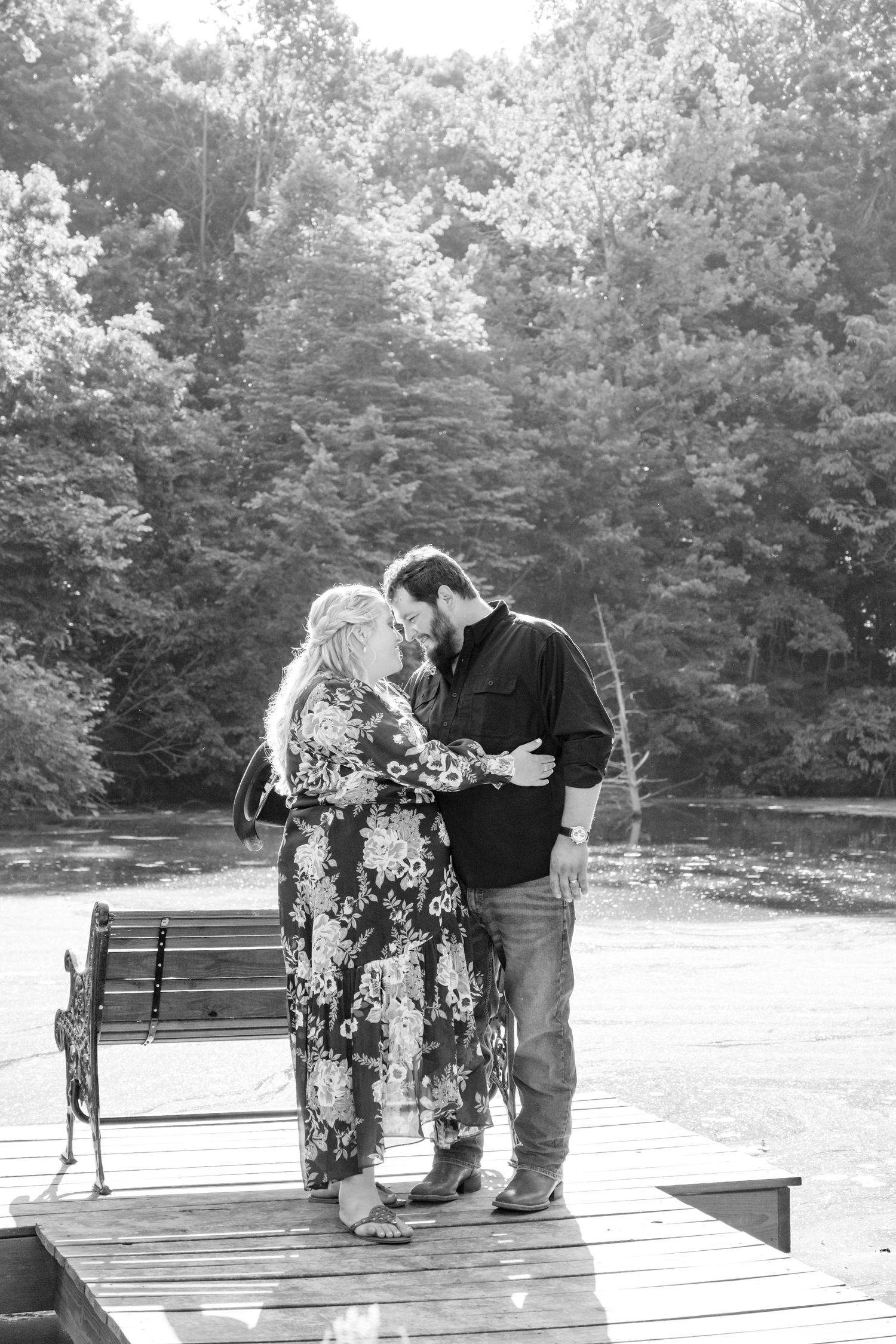 A couple stands together intimately on a wooden dock surrounded by trees in a black and white photograph.