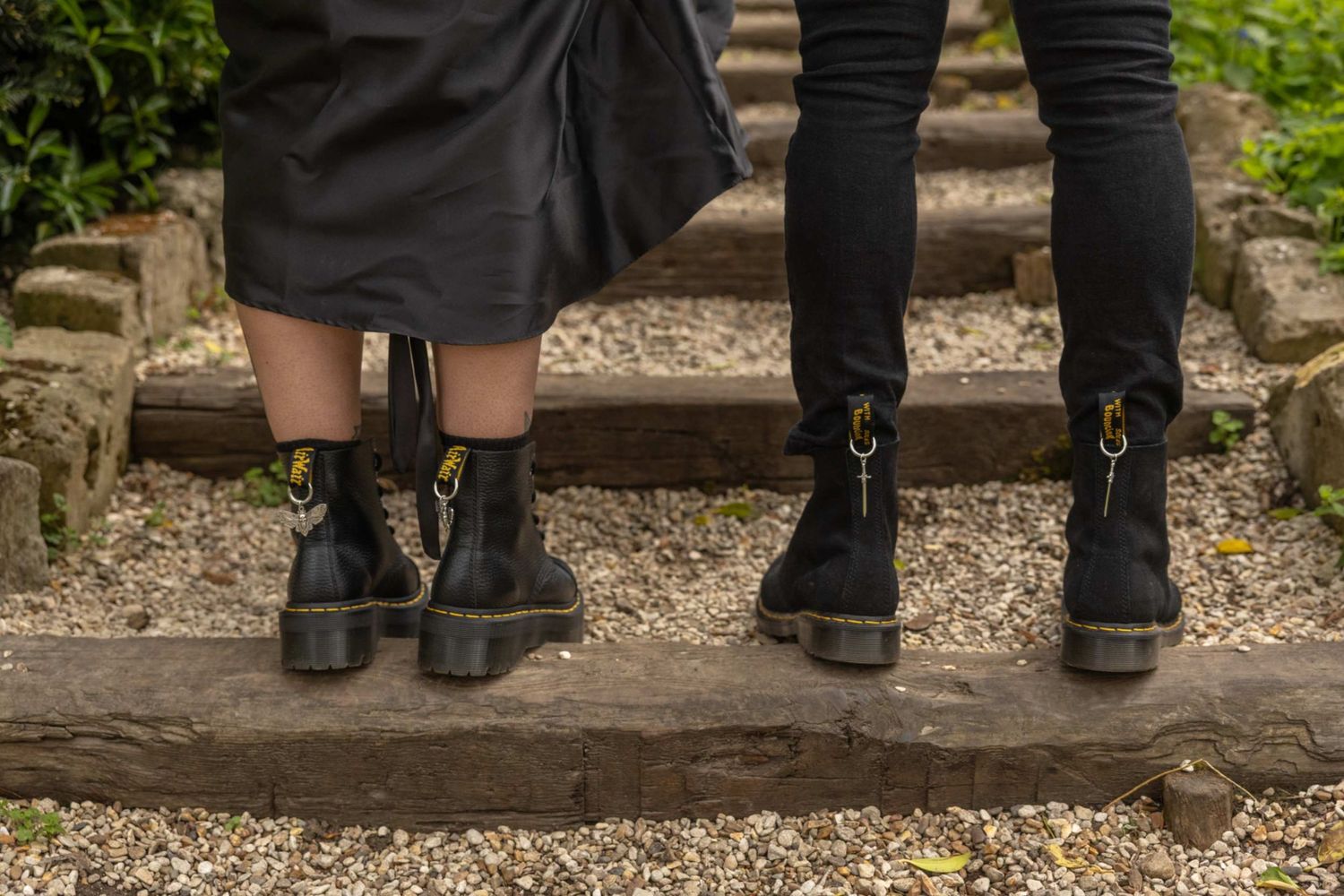 Two people wearing matching black combat boots standing on rustic wooden steps in a garden setting.