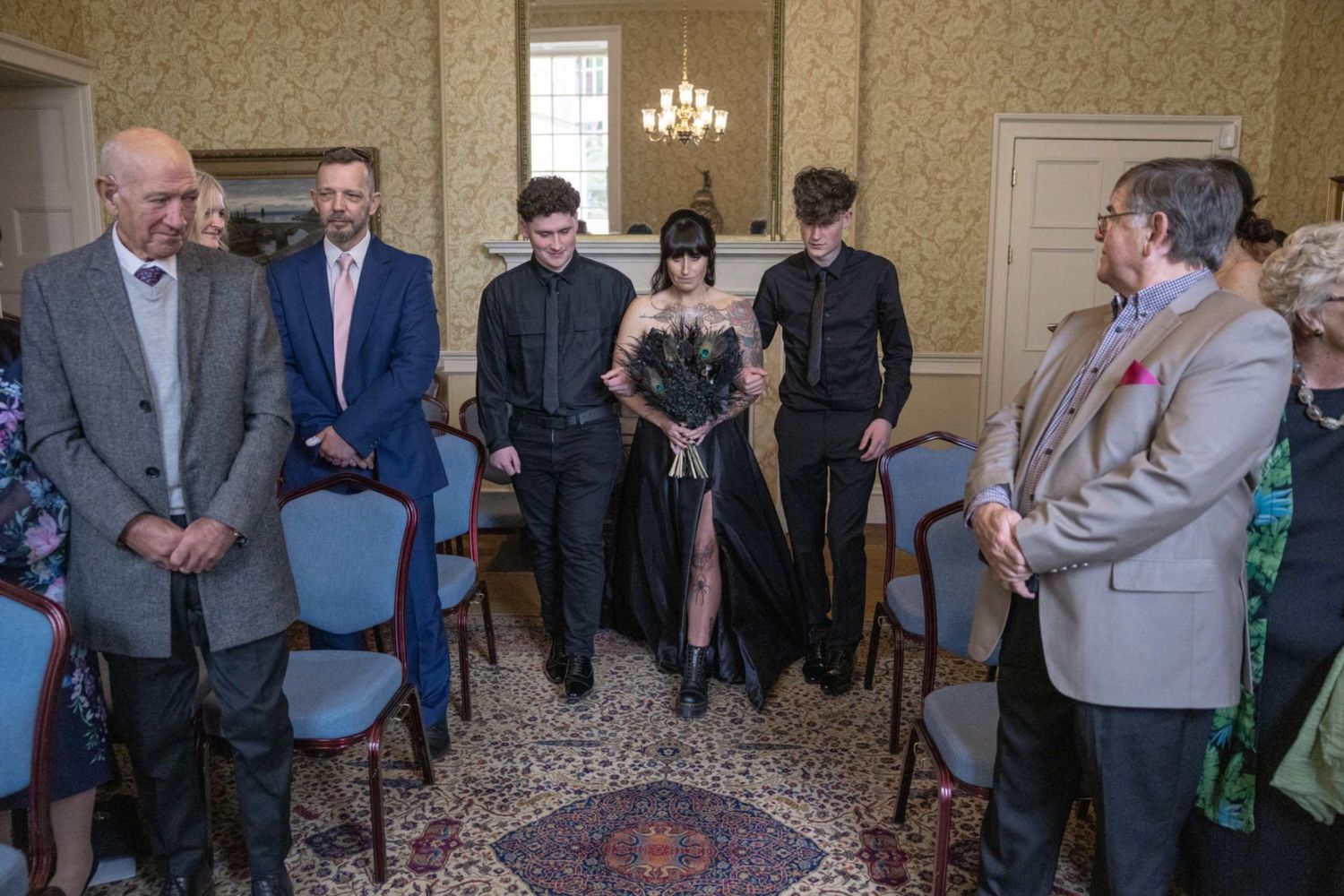 Small group of people gathered in a formal room with ornate wallpaper and a patterned rug for an indoor ceremony.