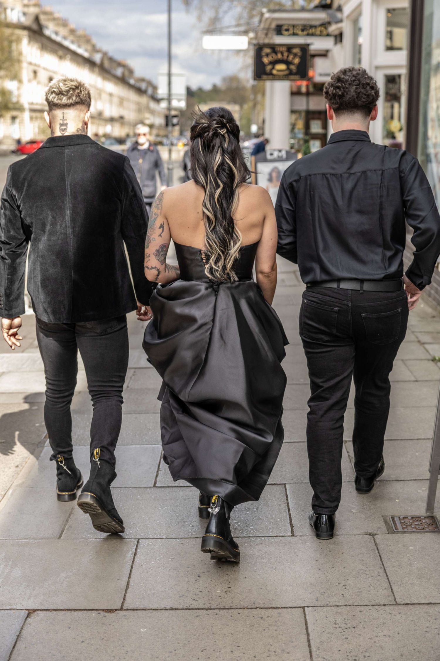 People in black attire walking on city sidewalk with woman in dark elegant dress with bow detail in the center.