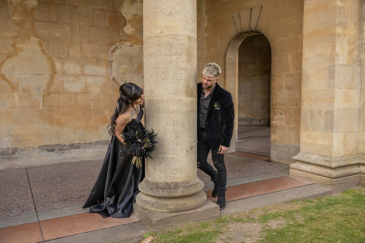 Gothic couple dressed in all black poses dramatically between stone pillars of historic building exterior.