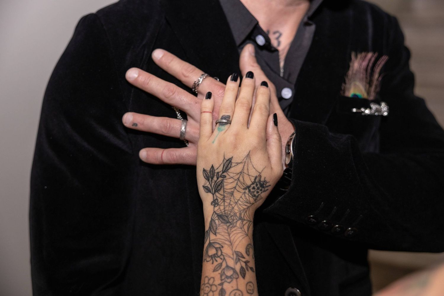 A hand with delicate floral tattoos showcases an engagement ring against a black velvet blazer background.