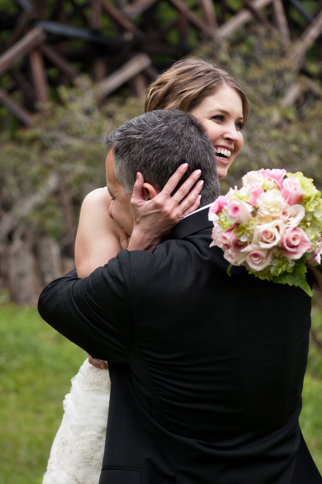 A joyful wedding embrace between newlyweds outdoors, with the bride holding a pink and white rose bouquet.