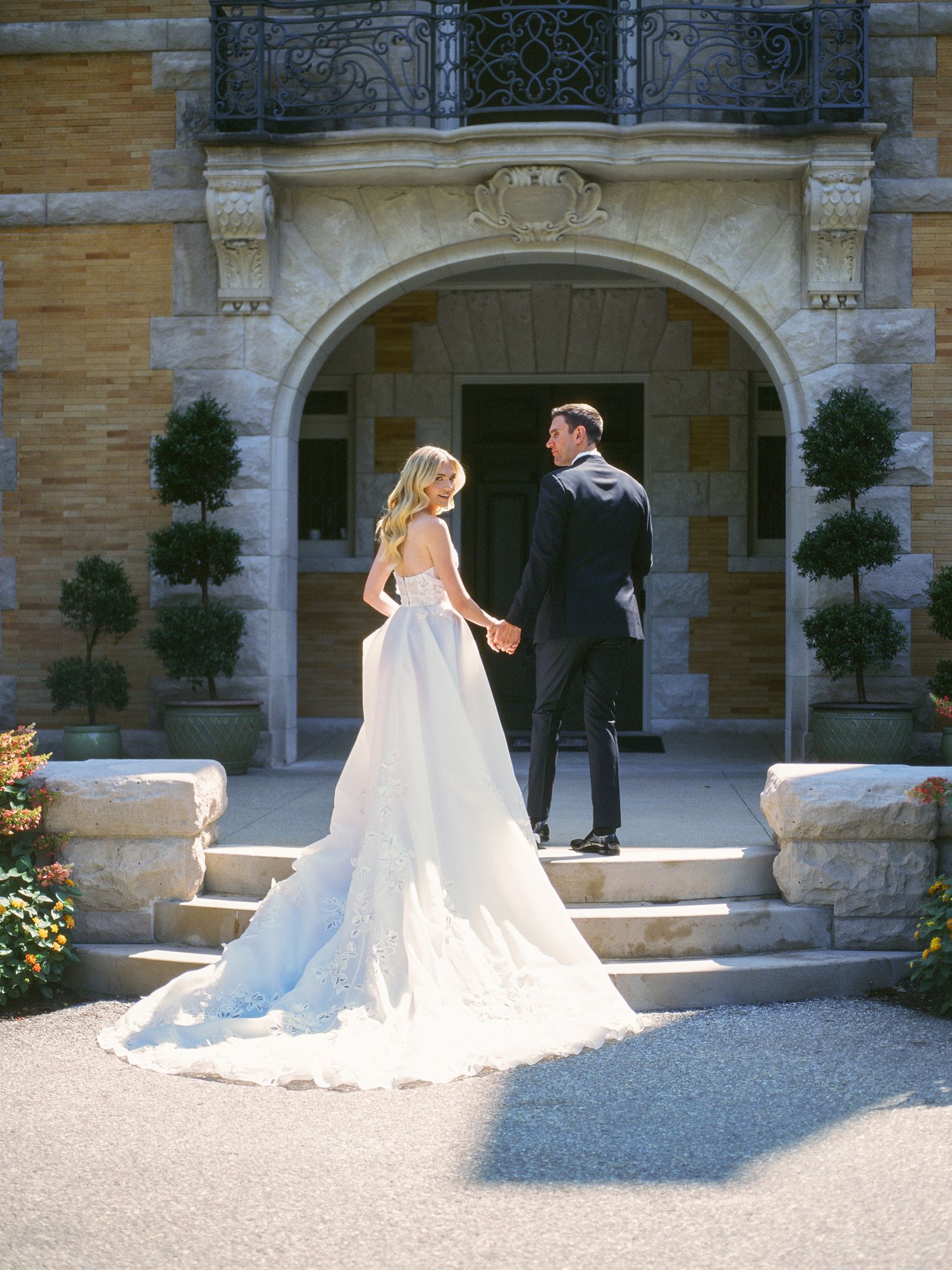 A bride in a flowing white wedding dress poses elegantly on a gravel path in front of a grand stone archway.