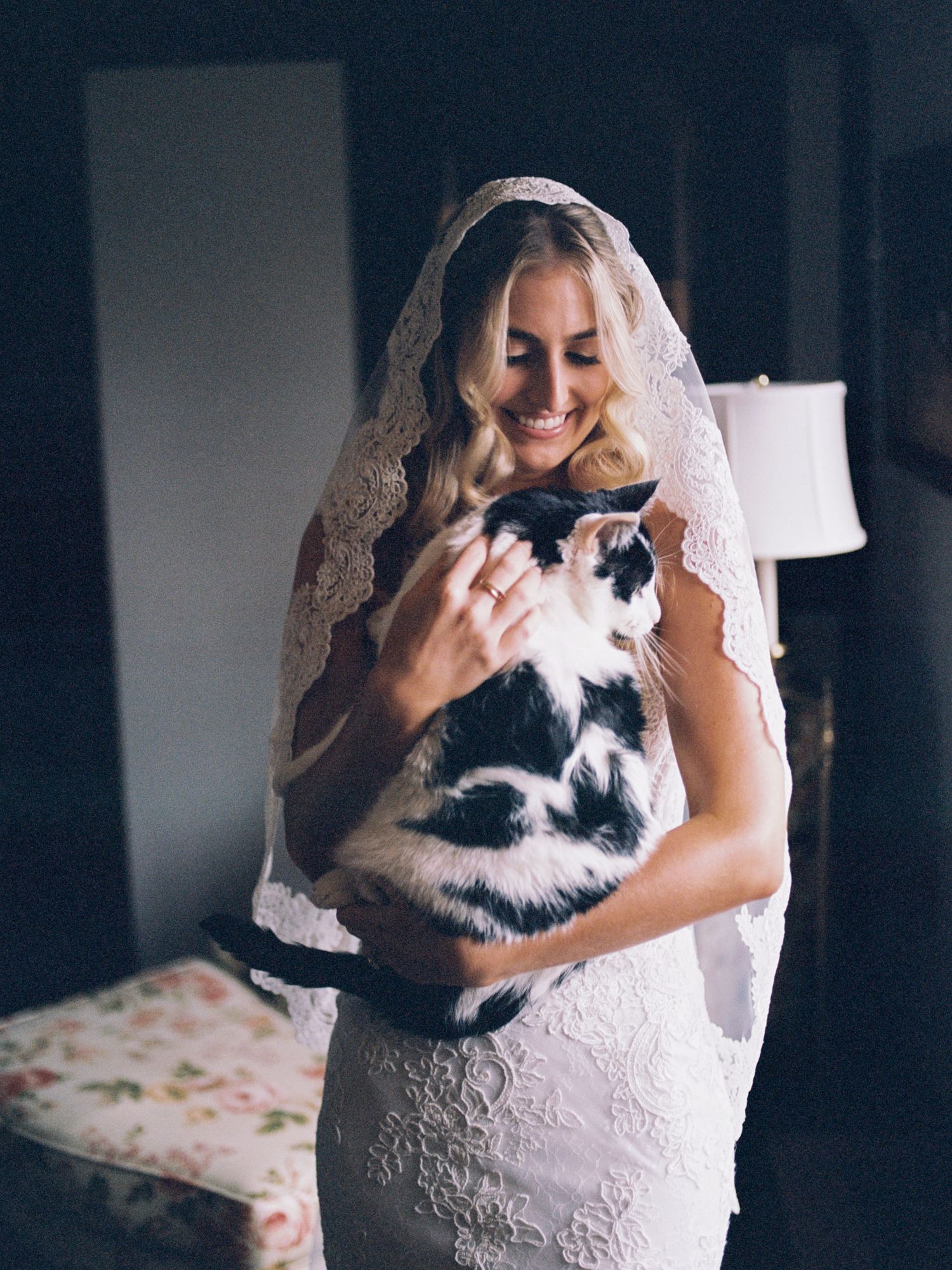 A bride in a white lace dress hugs her black and white cat while sitting on a floral bedspread.